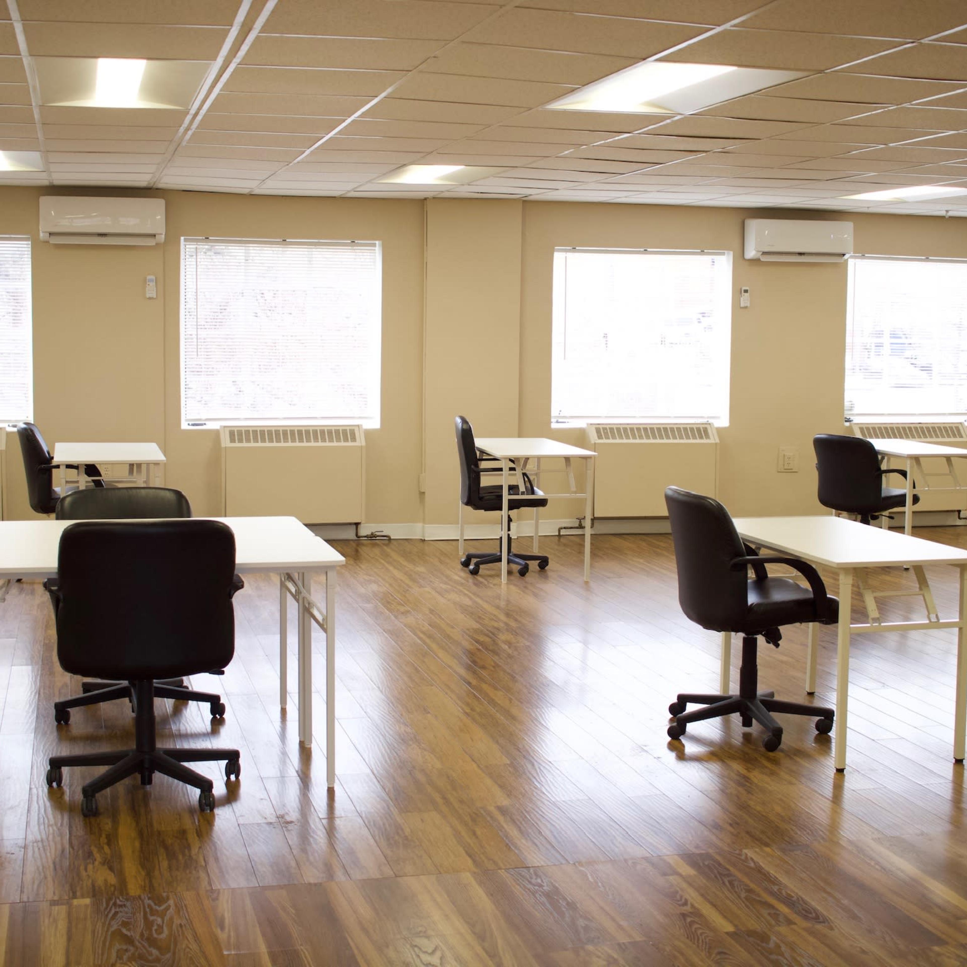 A spacious room with multiple desks and chairs arranged in a well-lit area near large windows.