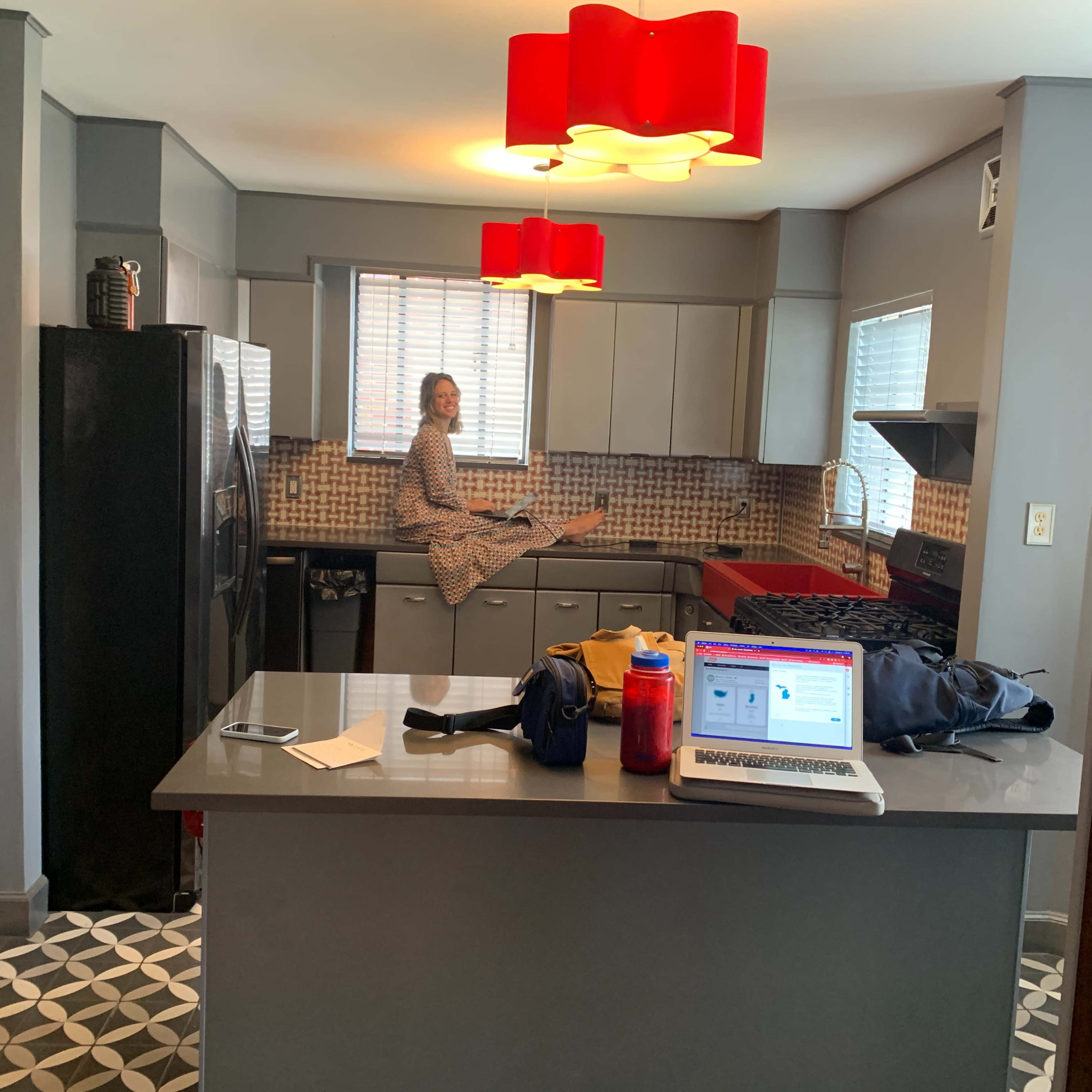 A person sits on a kitchen countertop in a modern kitchen with red pendant lights and a patterned floor.