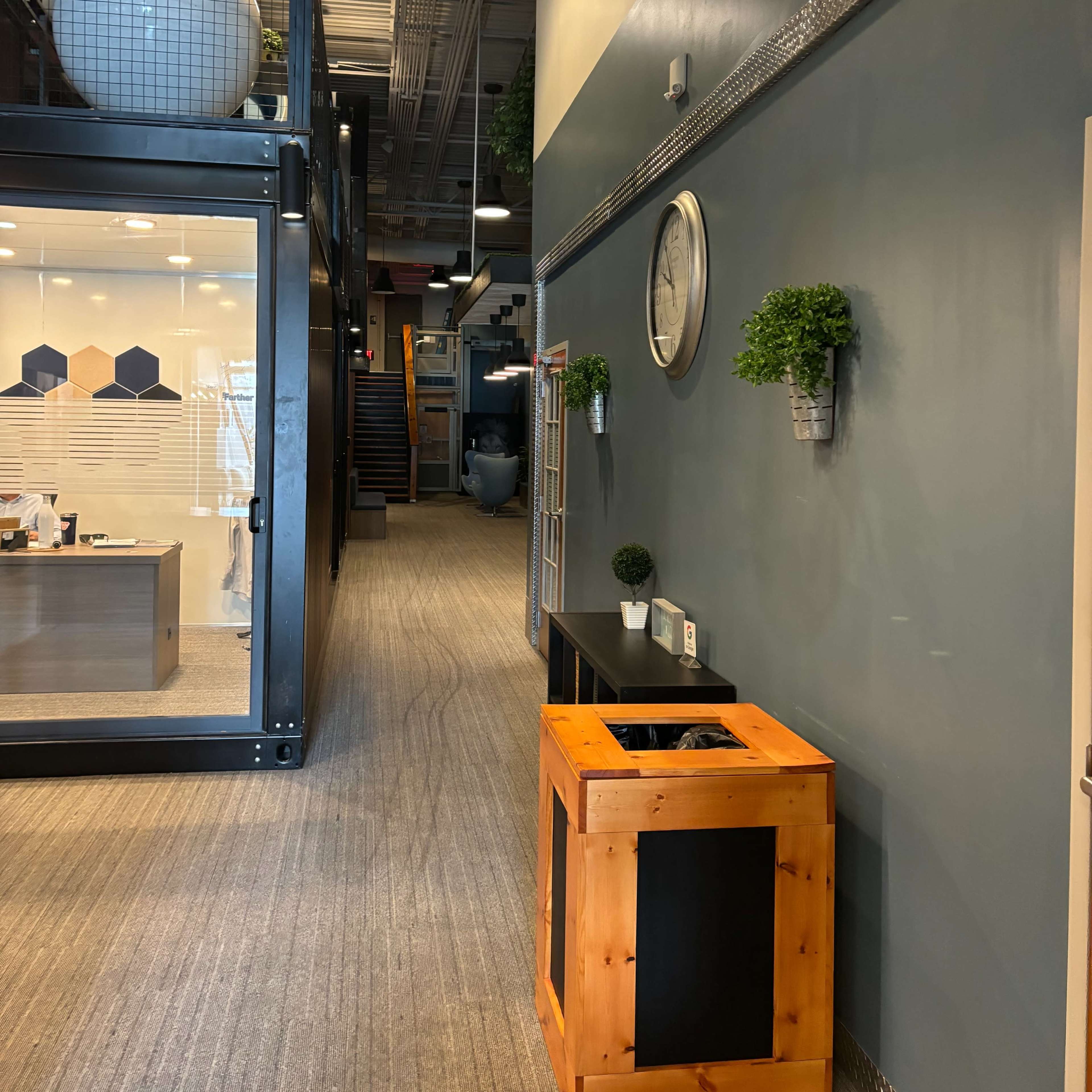 A modern office corridor with a wooden trash bin, decorative plants on the wall, and visible staircases in the background.
