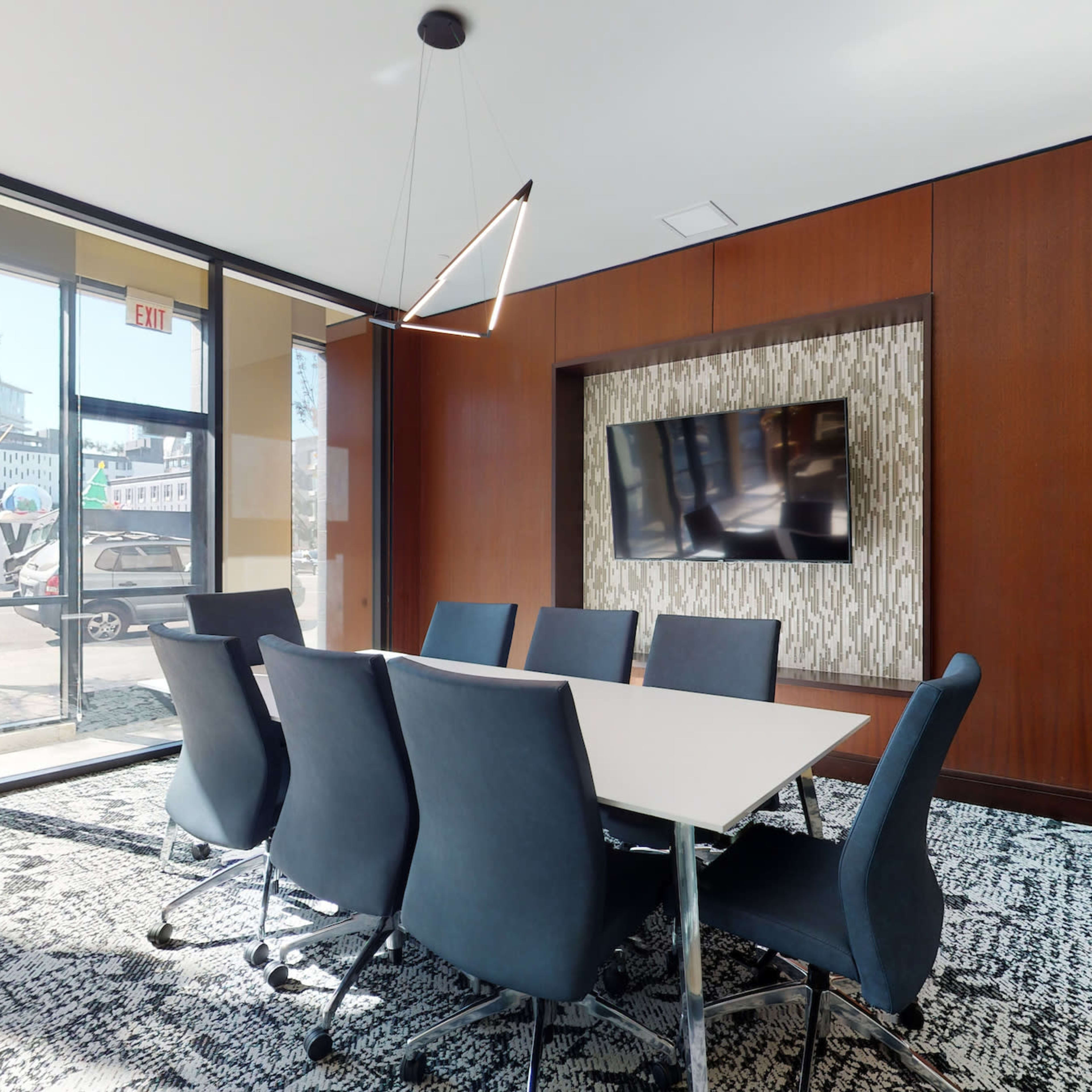 A modern conference room features a large table surrounded by ergonomic chairs, a wall-mounted TV, and large windows that provide natural light.