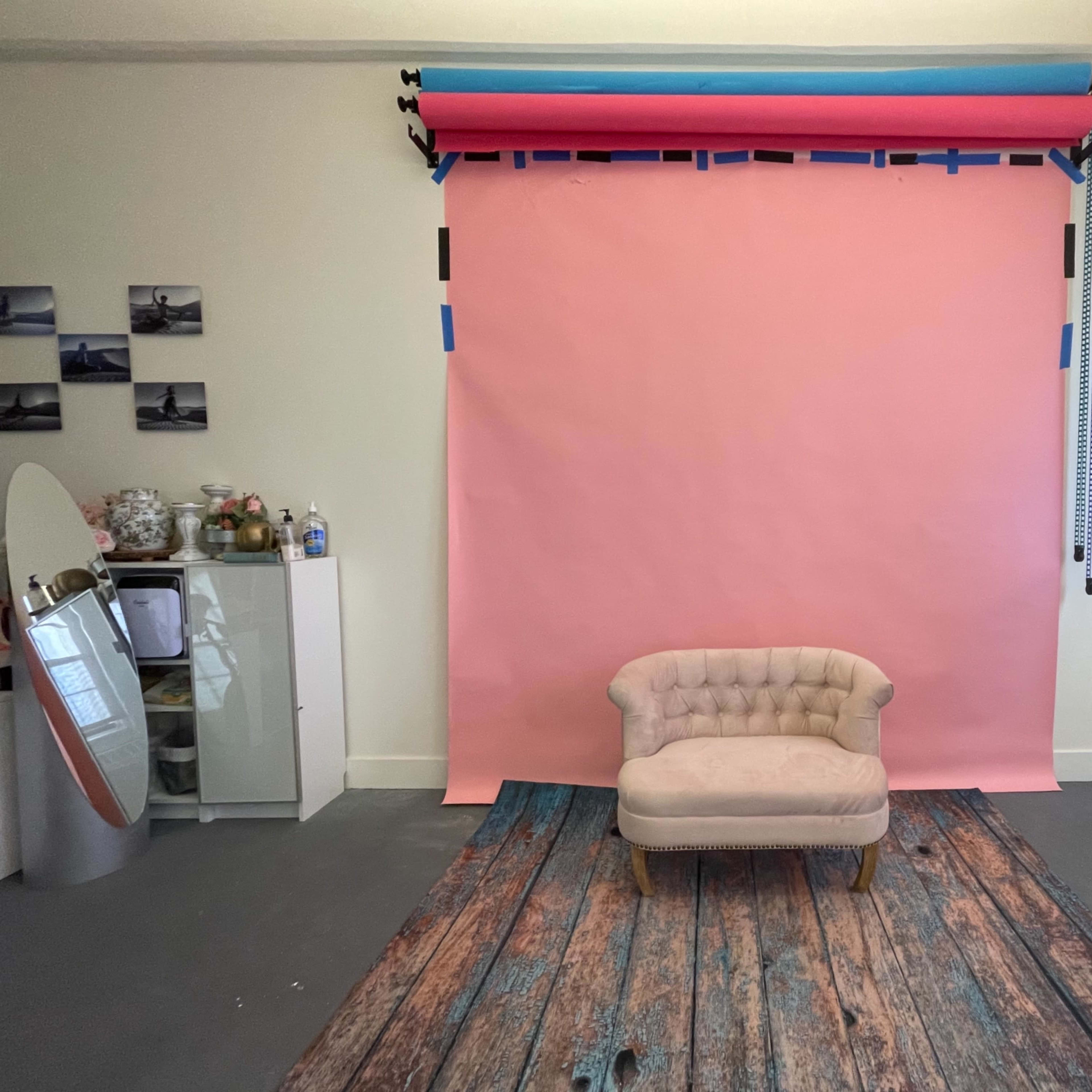 A simple studio setup with a pink backdrop, a wooden floor, and a tufted beige chair in front.