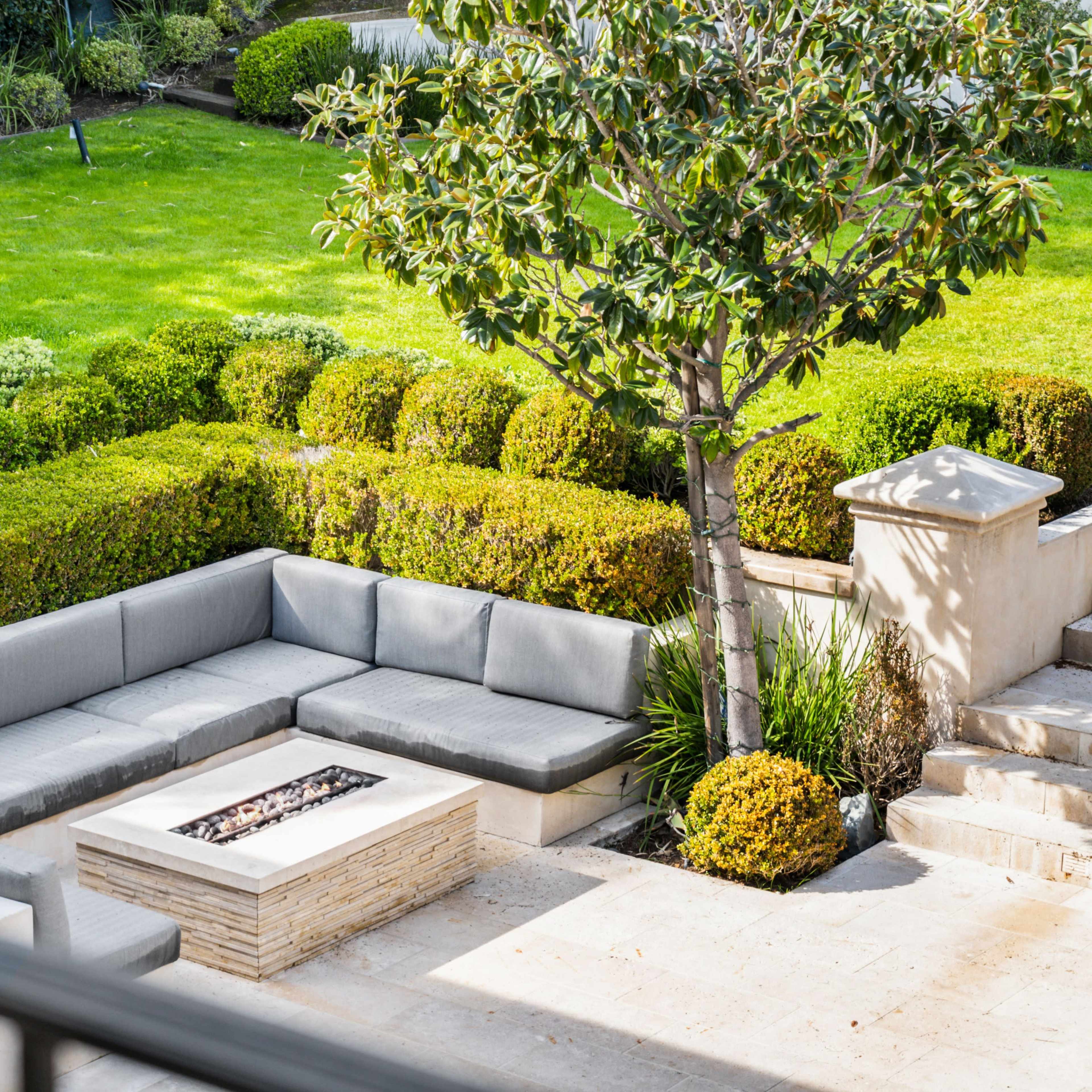 A backyard equipped with a gray sectional sofa surrounding a stone fire pit, nestled among well-maintained greenery and landscaping.
