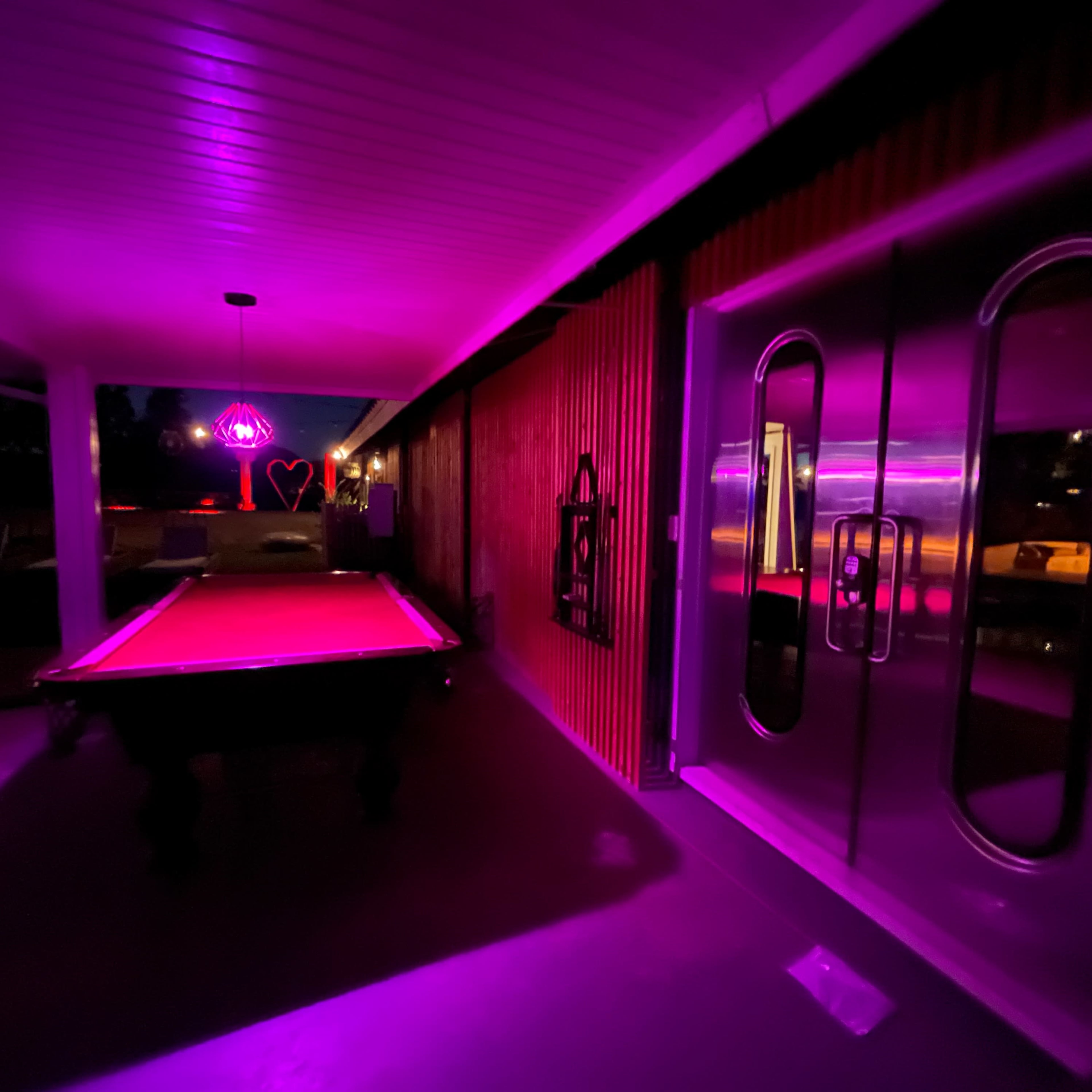 A dimly lit game room features a pool table under purple lighting, with large glass doors reflecting the vibrant colors.