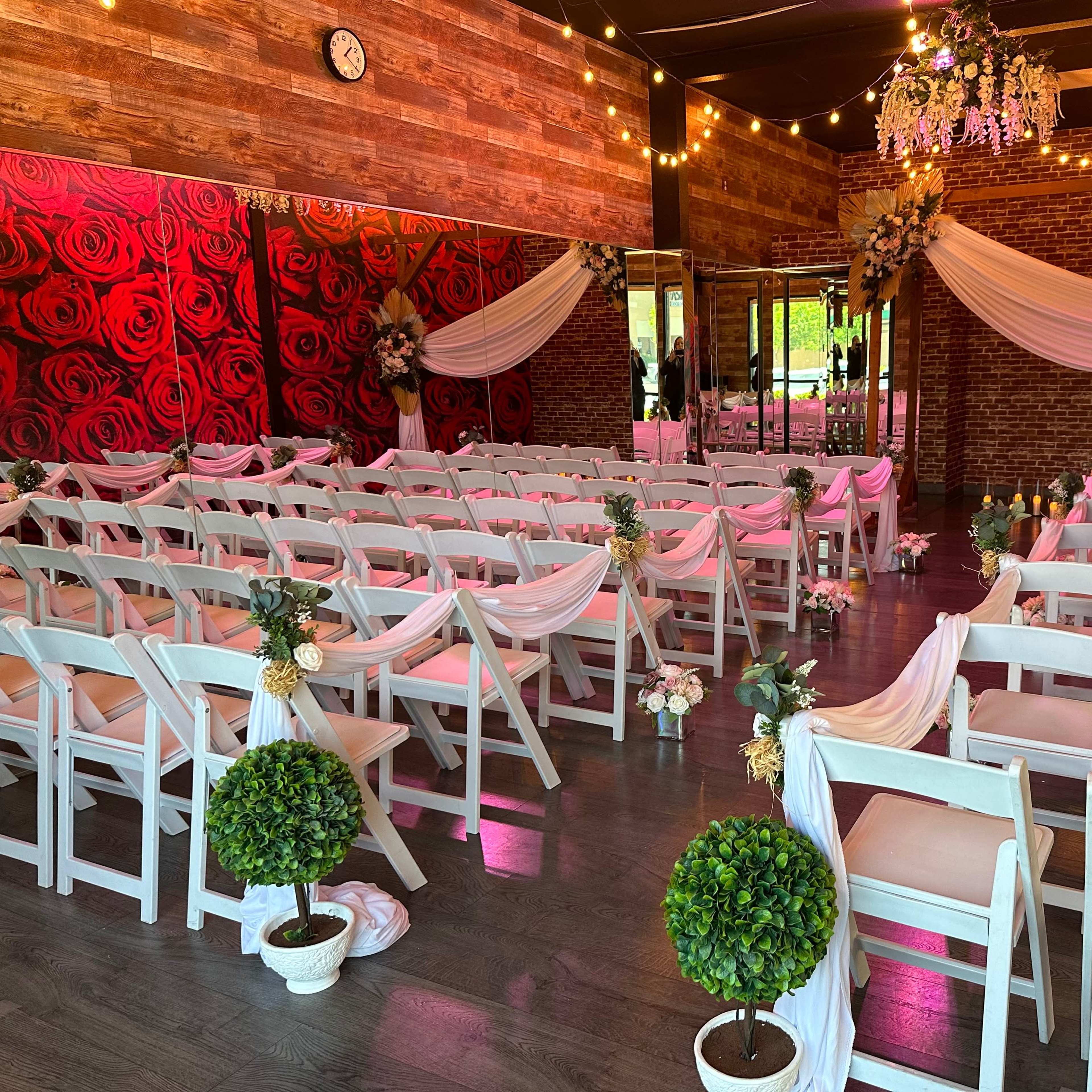 A decorated indoor space set up for a wedding ceremony, featuring rows of white chairs adorned with floral accents and draped fabric, surrounded by wooden and floral decor.