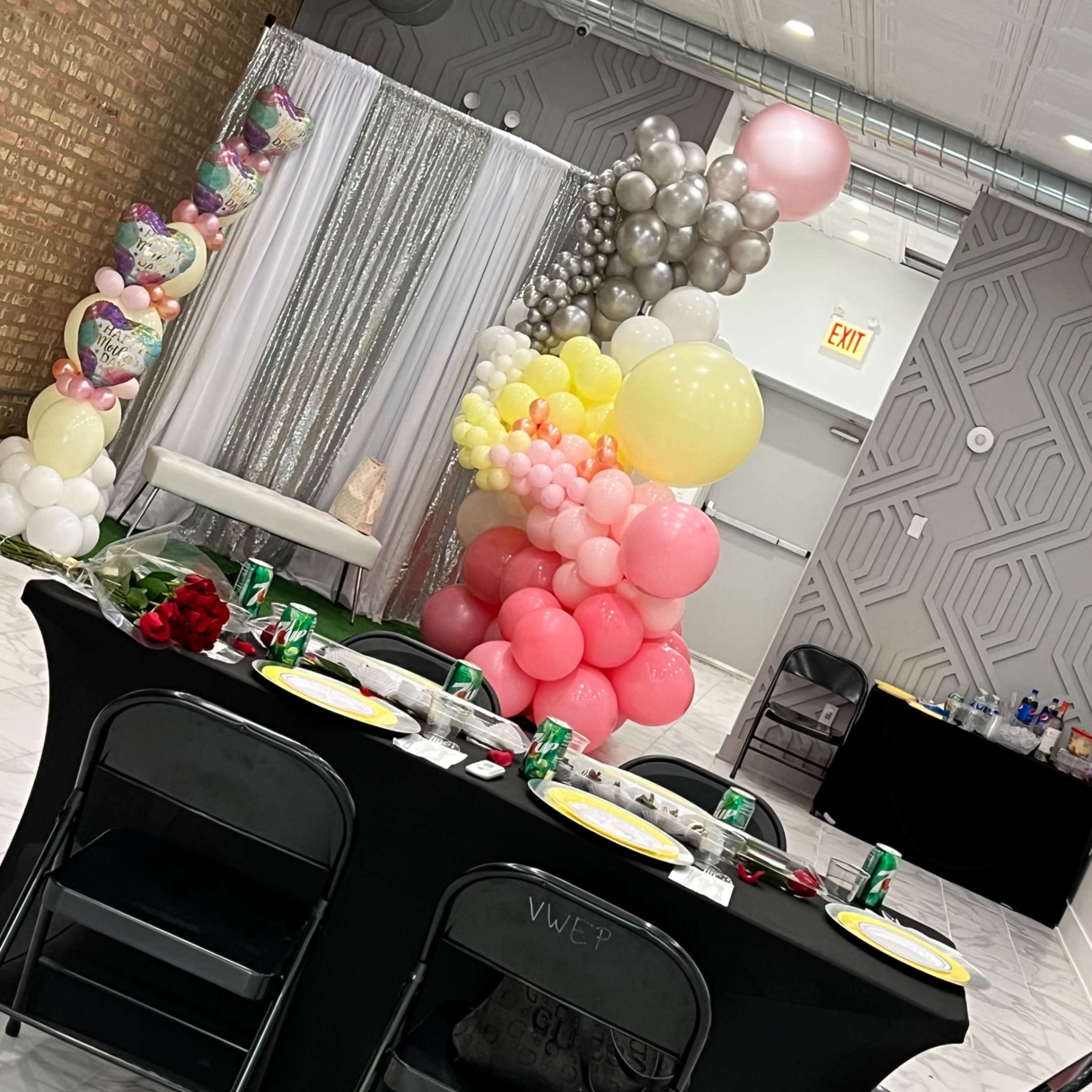 A decorated event space features a table set with plates and drinks, surrounded by a backdrop of balloons and a shimmering curtain.