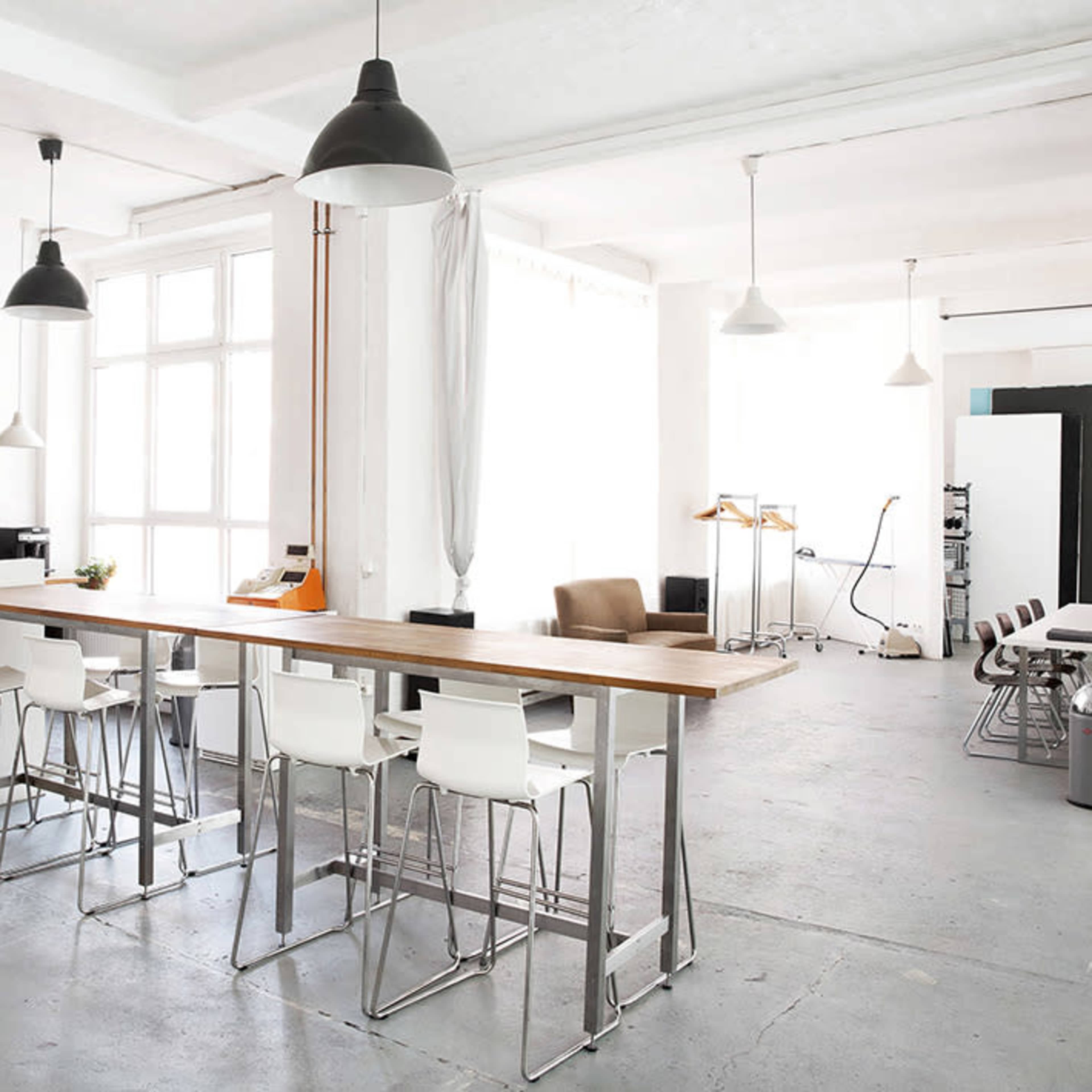 A spacious, modern kitchen area with a long wooden bar and white stools, surrounded by bright walls and industrial lighting.