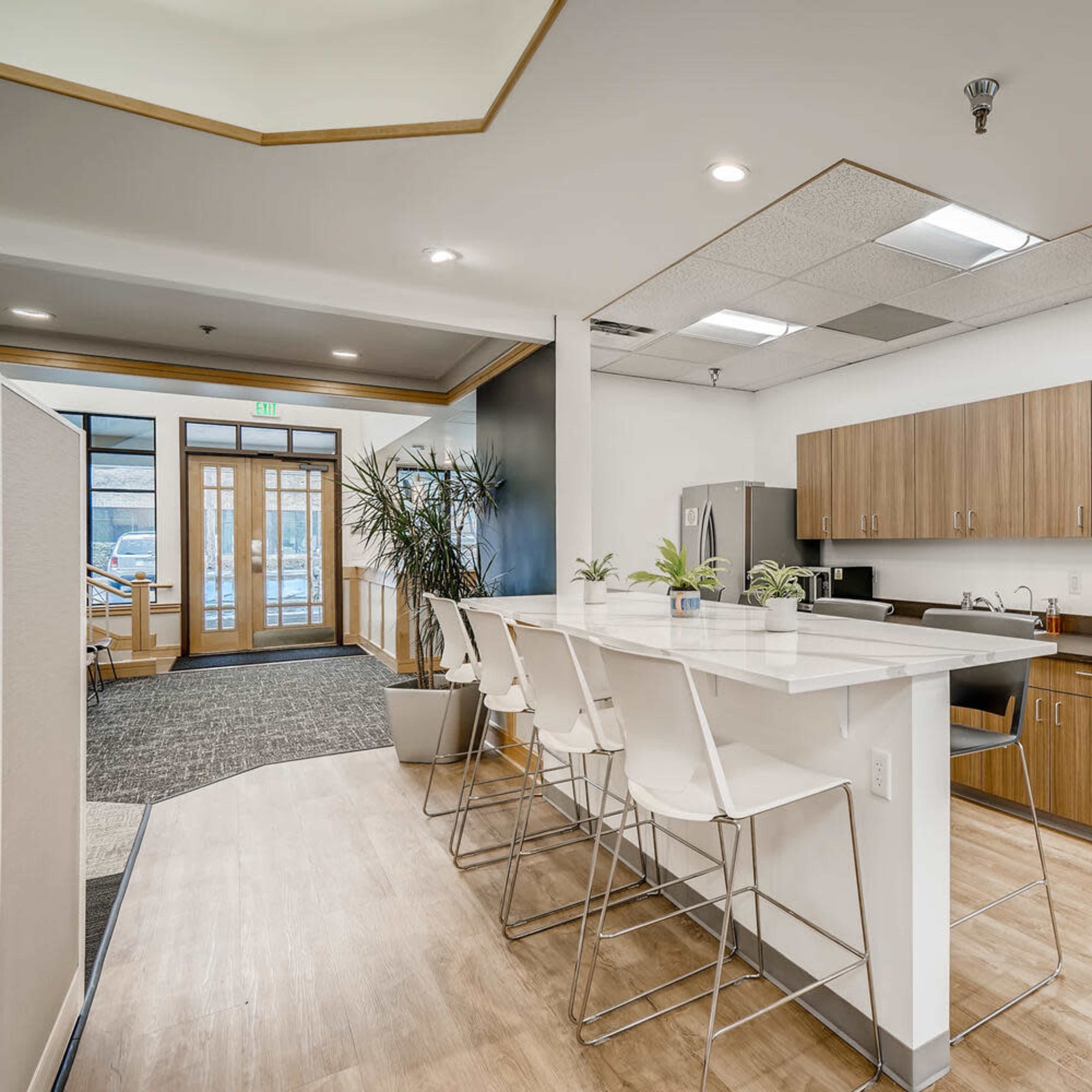 A modern kitchen area with wooden cabinets, a white countertop, and high stools, surrounded by potted plants.