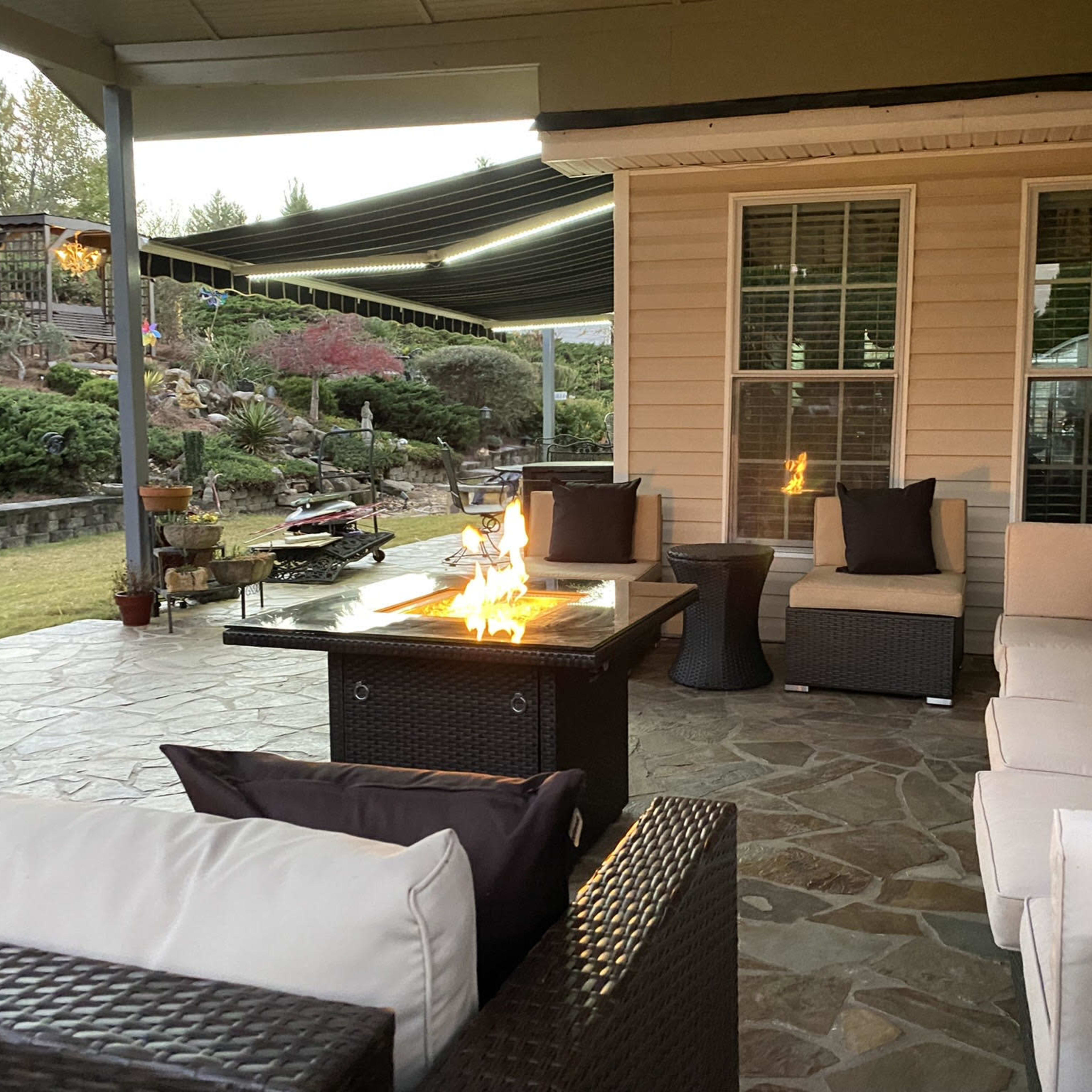 A patio area featuring a fire pit table surrounded by outdoor seating and a landscaped yard in the background.