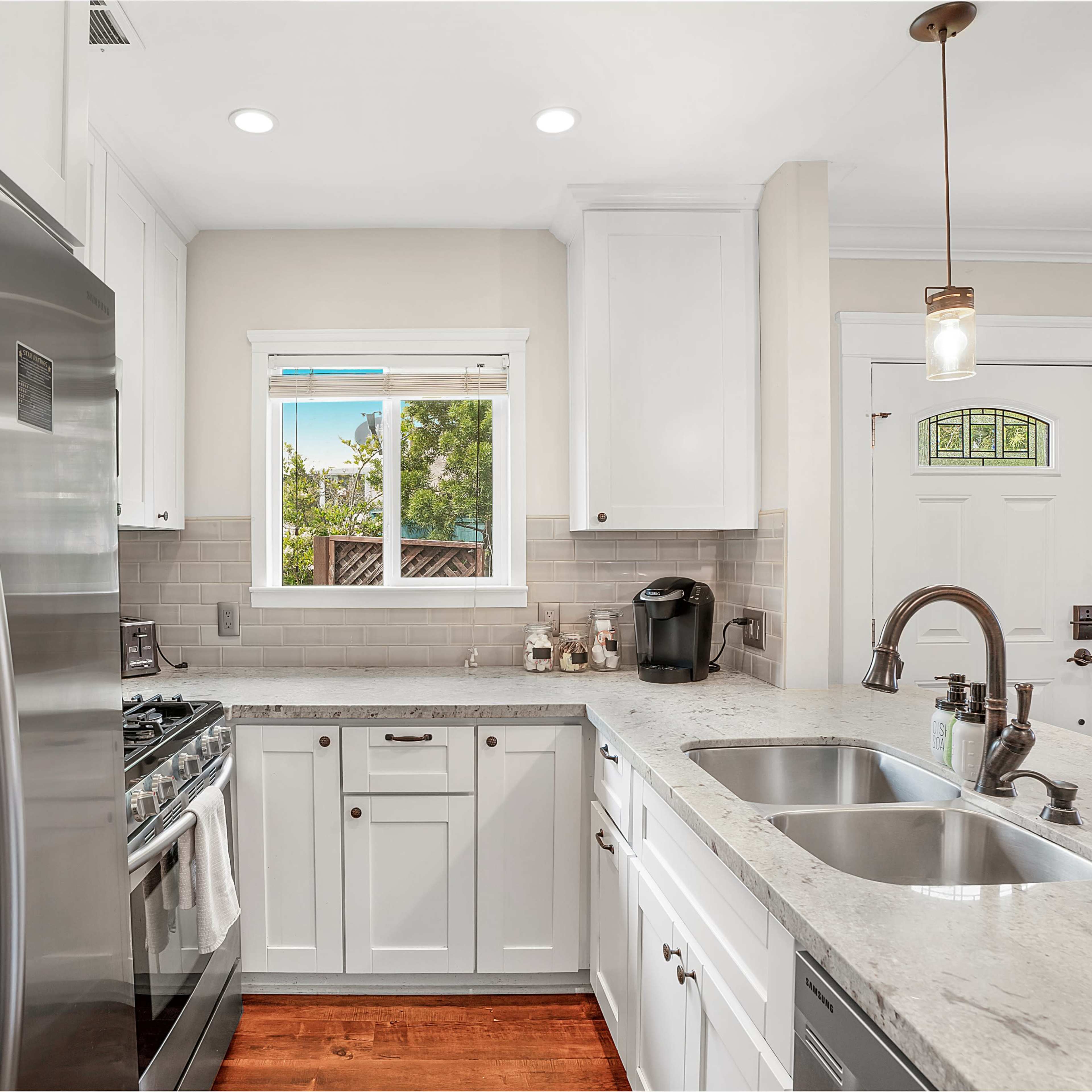 A modern kitchen features stainless steel appliances, white cabinets, a double sink, and light-colored countertops with a window overlooking an outdoor area.