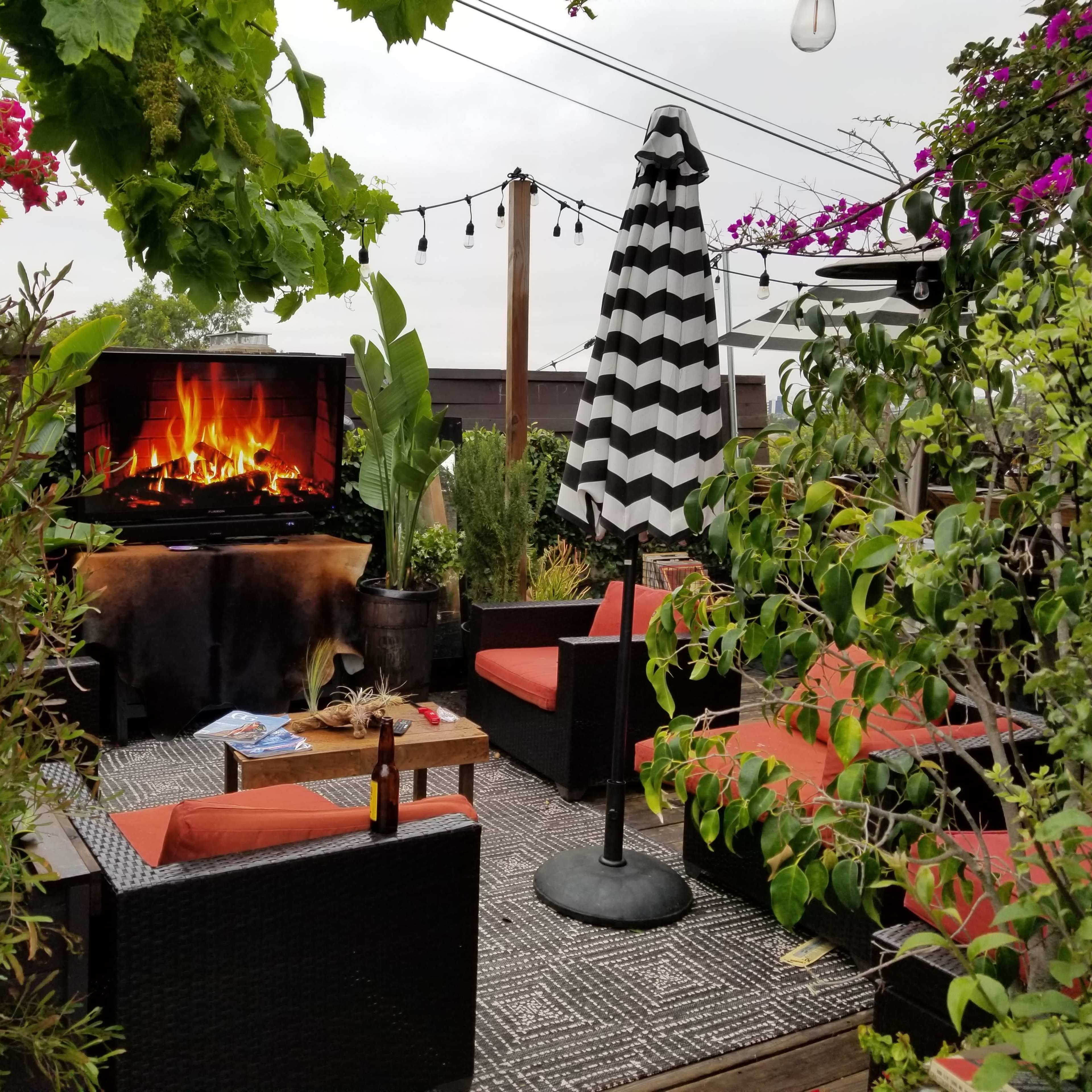 A cozy outdoor seating area features a faux fireplace, surrounded by greenery and decorative plants, with a striped umbrella and lounge chairs.
