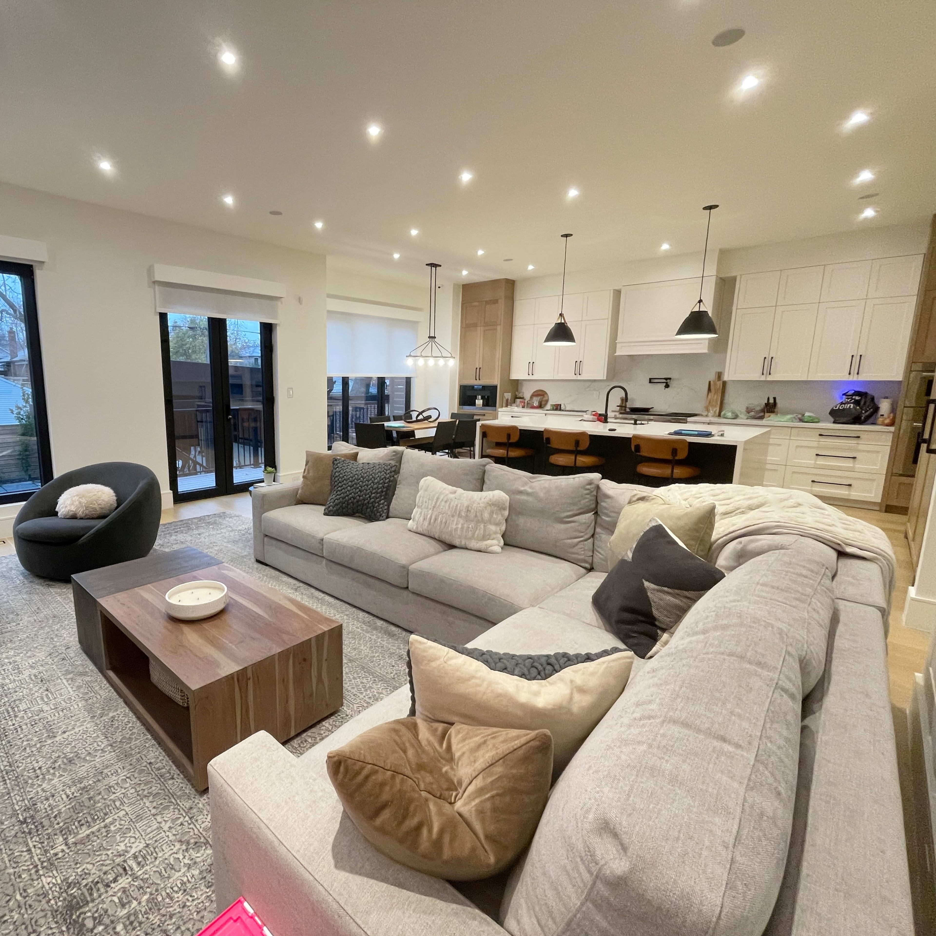 A modern, open-concept living area featuring a large sectional sofa, a coffee table, and a view of a well-lit kitchen with island seating.