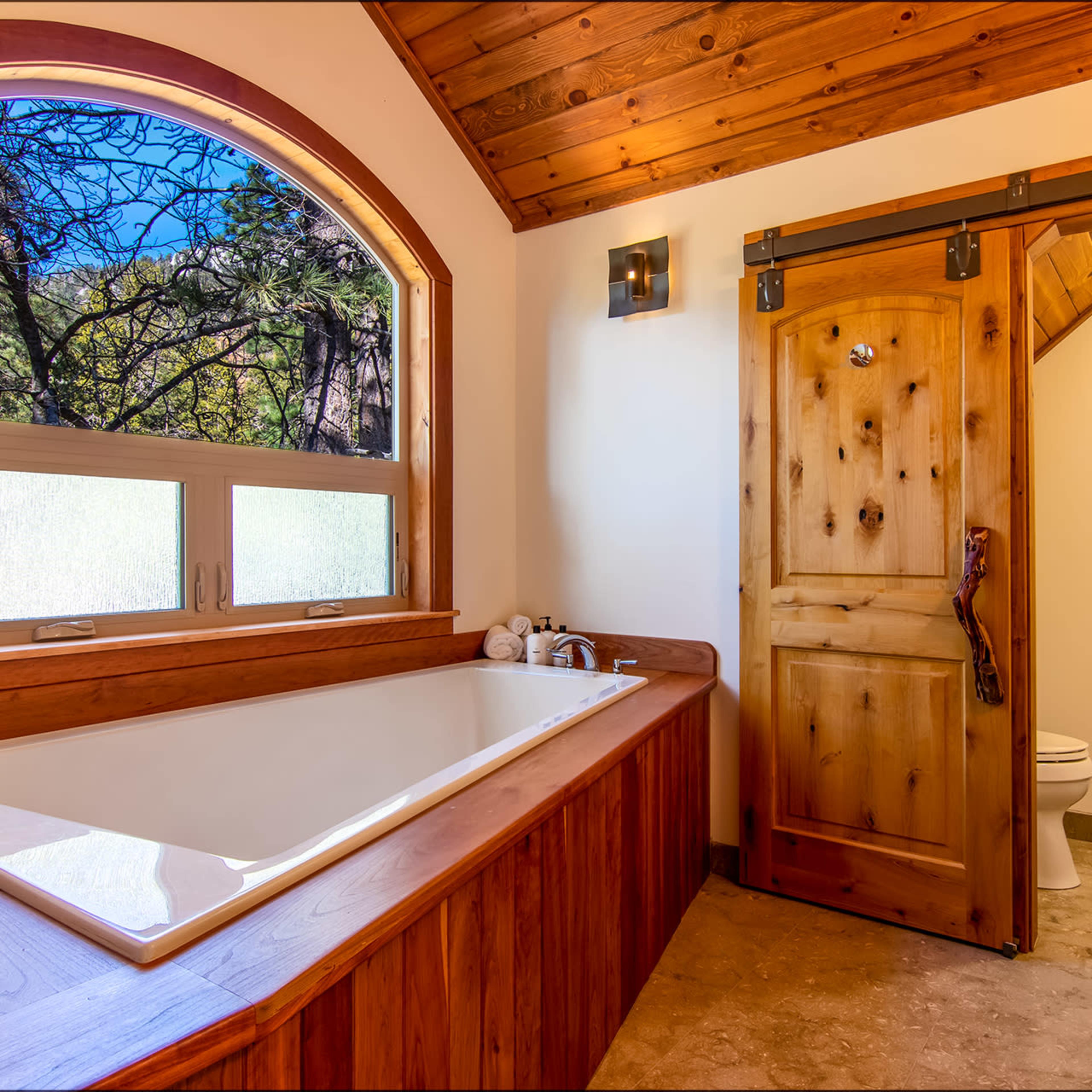 A wooden bathroom featuring a freestanding bathtub beside a large window and a sliding wooden door leading to a toilet area.