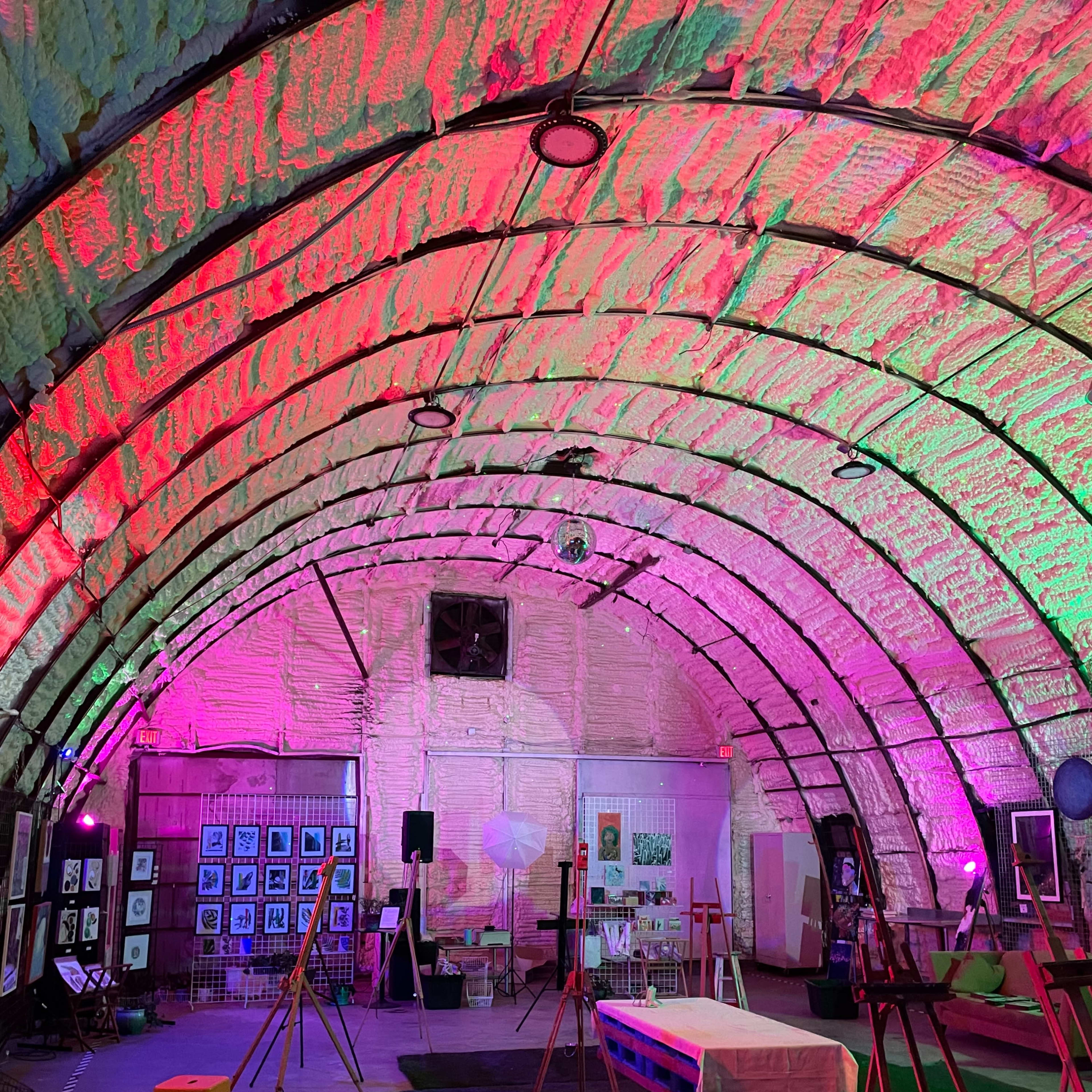 The interior of a large, dome-shaped space is illuminated with pink and green lighting, featuring art displays and setup for an event.
