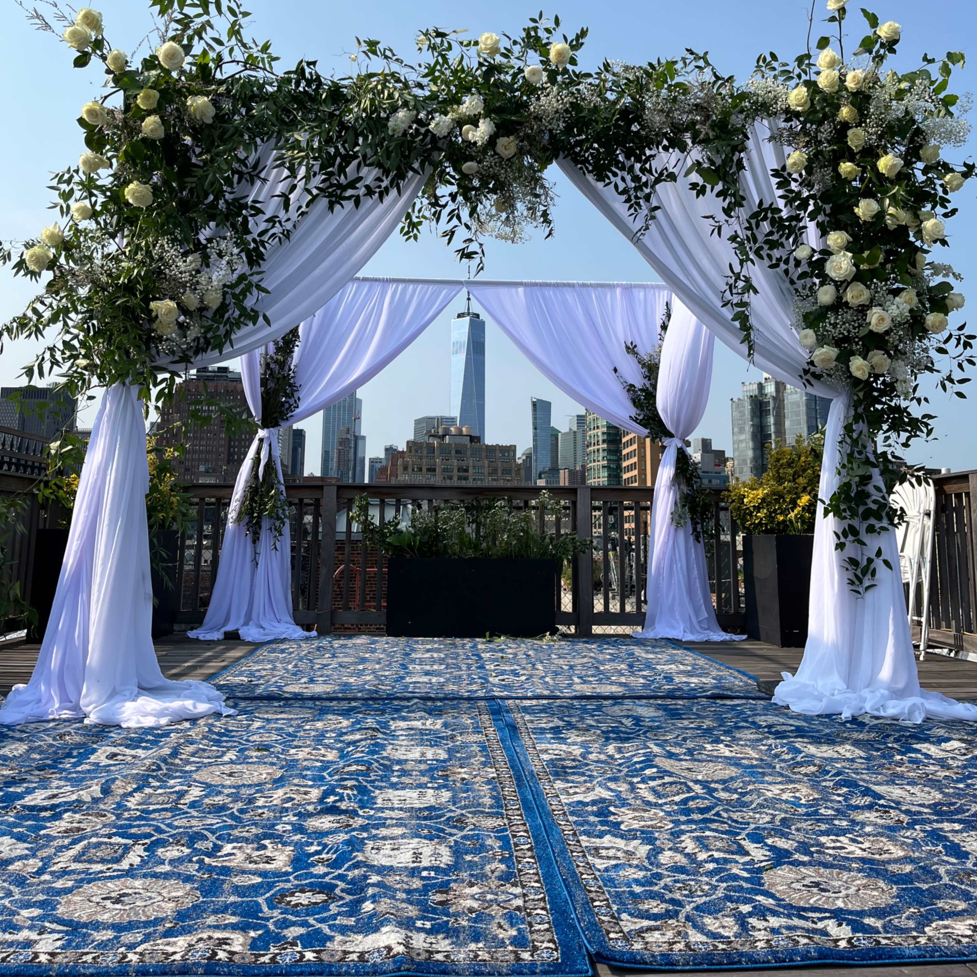 A wedding altar adorned with white drapes and floral arrangements, set against a skyline featuring a tall building in the background.
