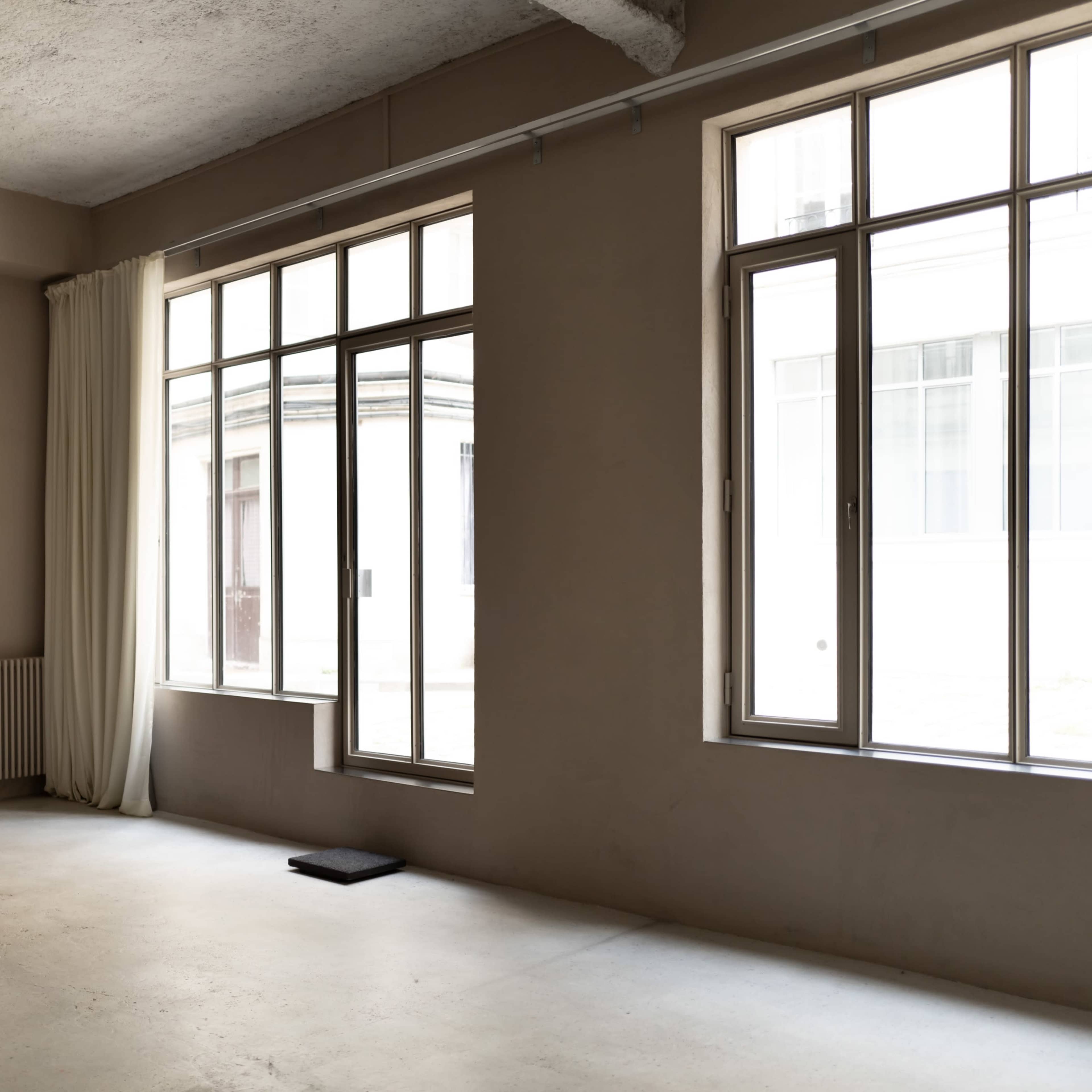An empty interior space features large windows, light-colored walls, and a radiator along one wall.