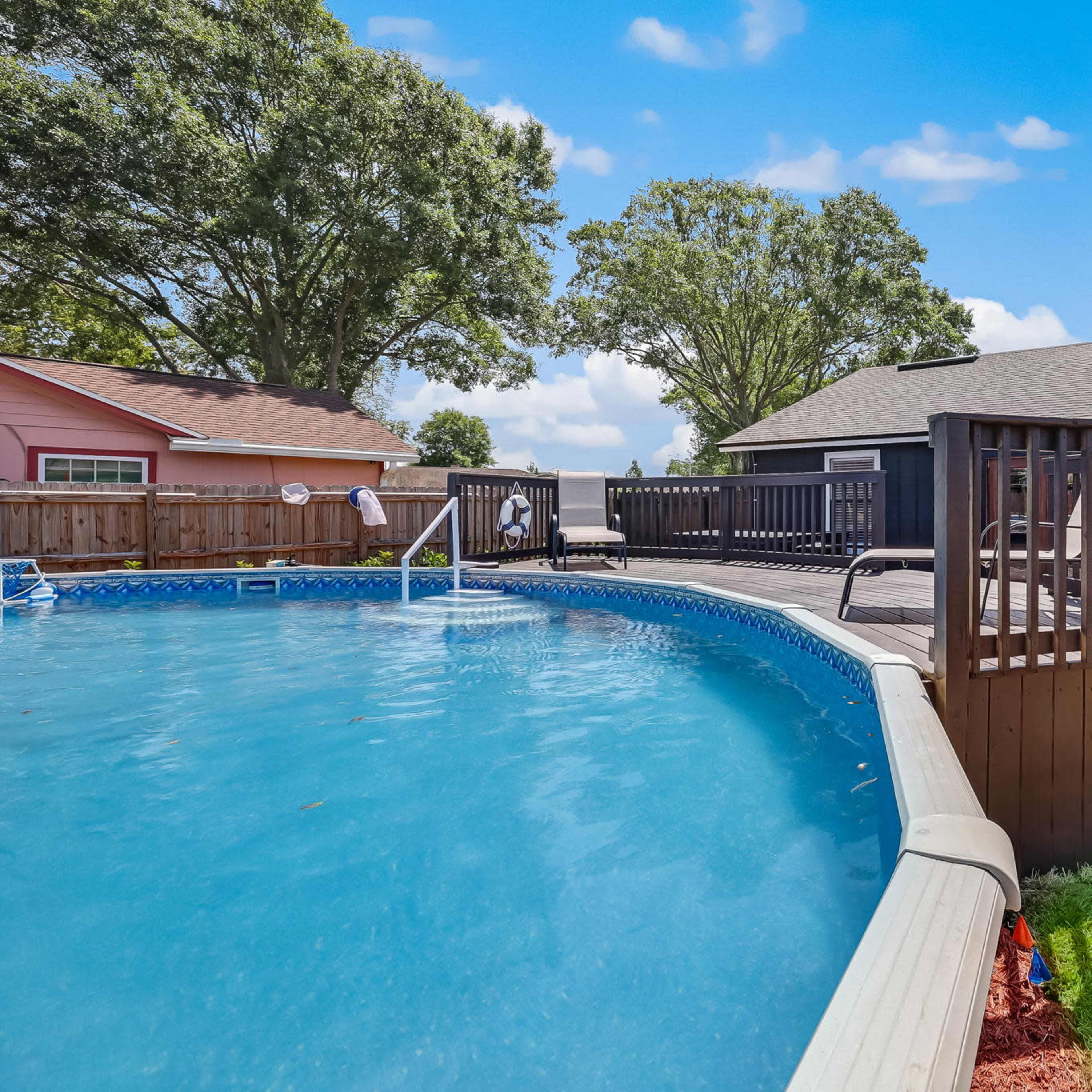 A circular above-ground pool surrounded by a wooden deck and lounge chairs, with trees in the background and a house nearby.