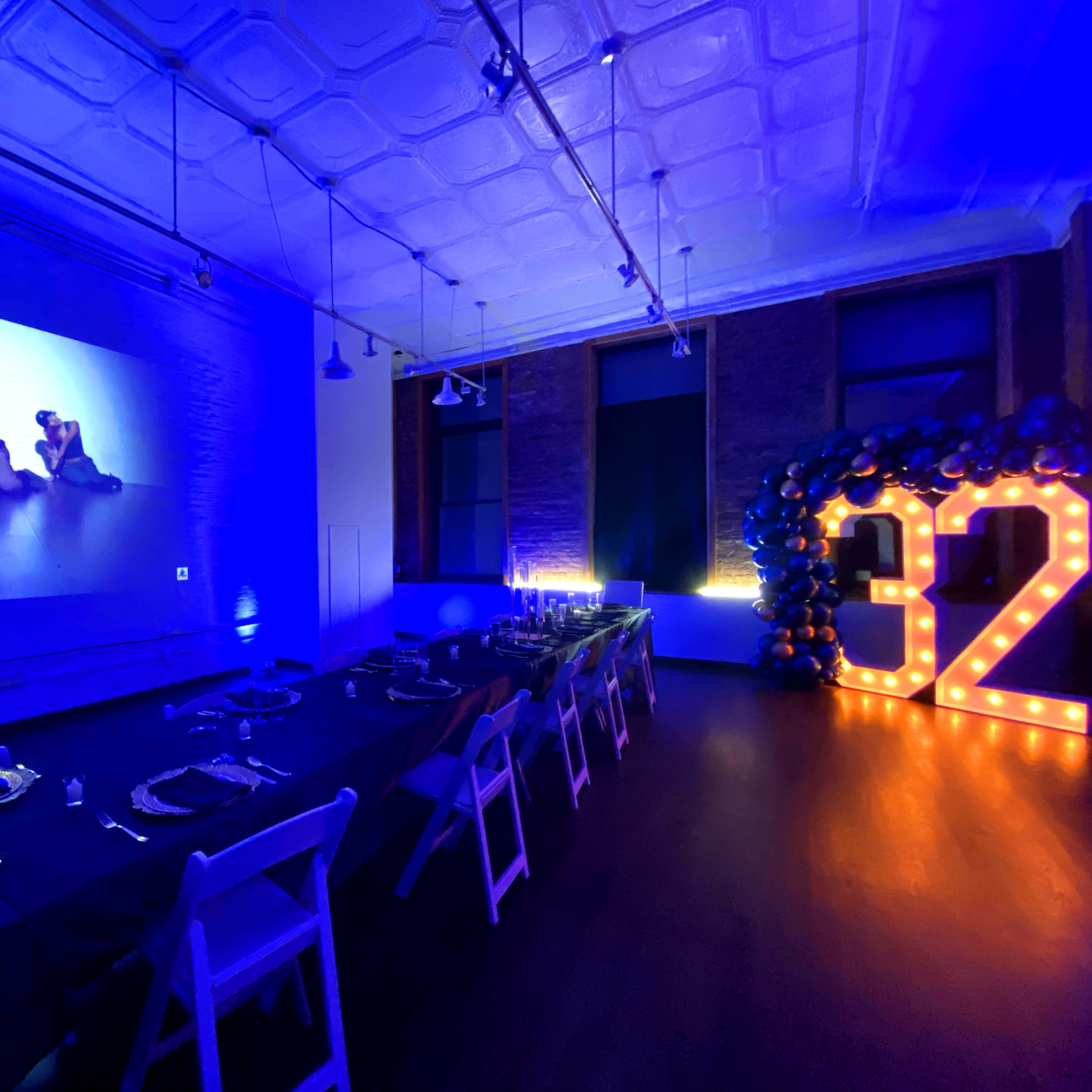 A dining area is set with a long table adorned with elegant place settings, illuminated by blue lighting and featuring a large illuminated "32" alongside a projector display.