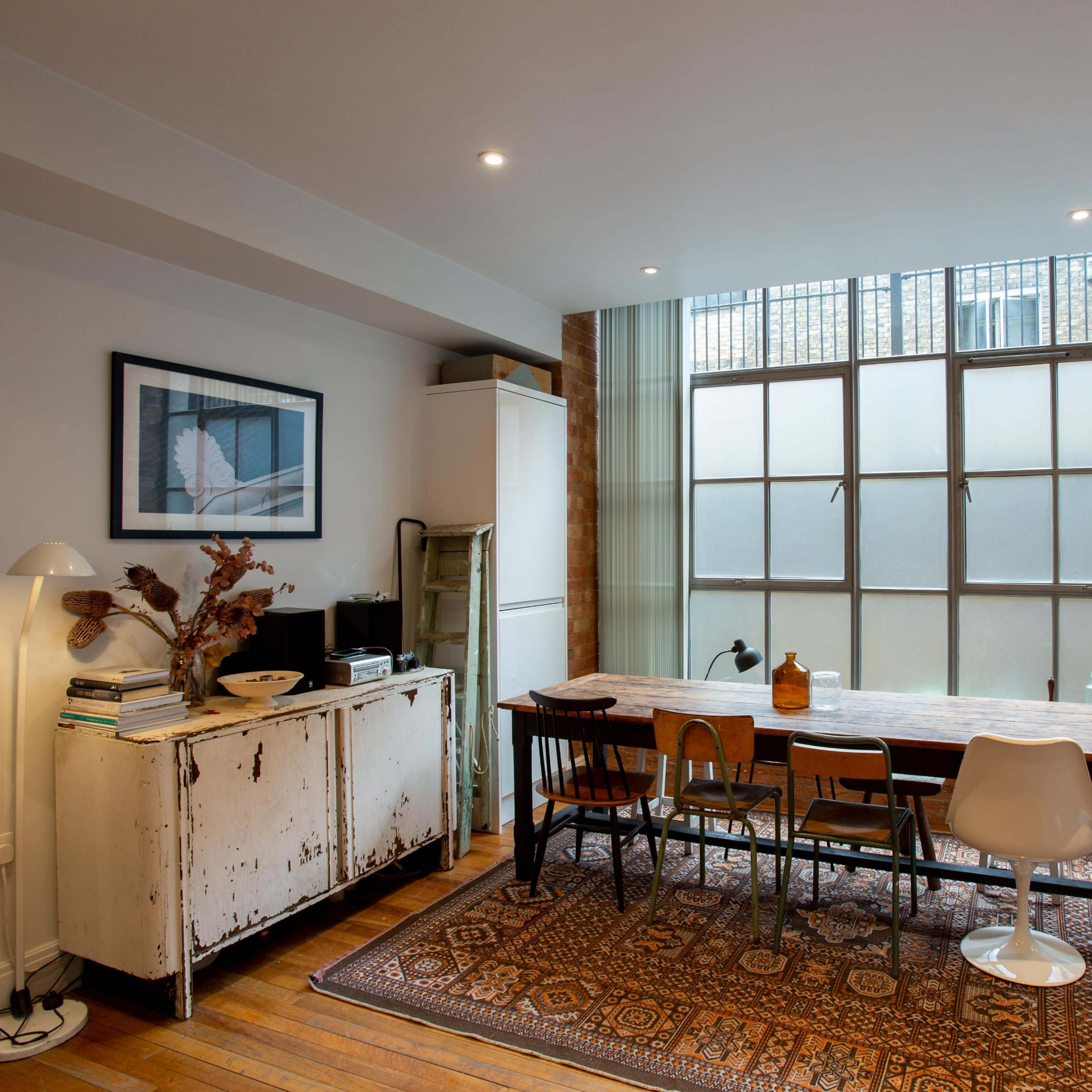 A bright dining area features a long wooden table surrounded by chairs, against a large window with a view of the outdoors.