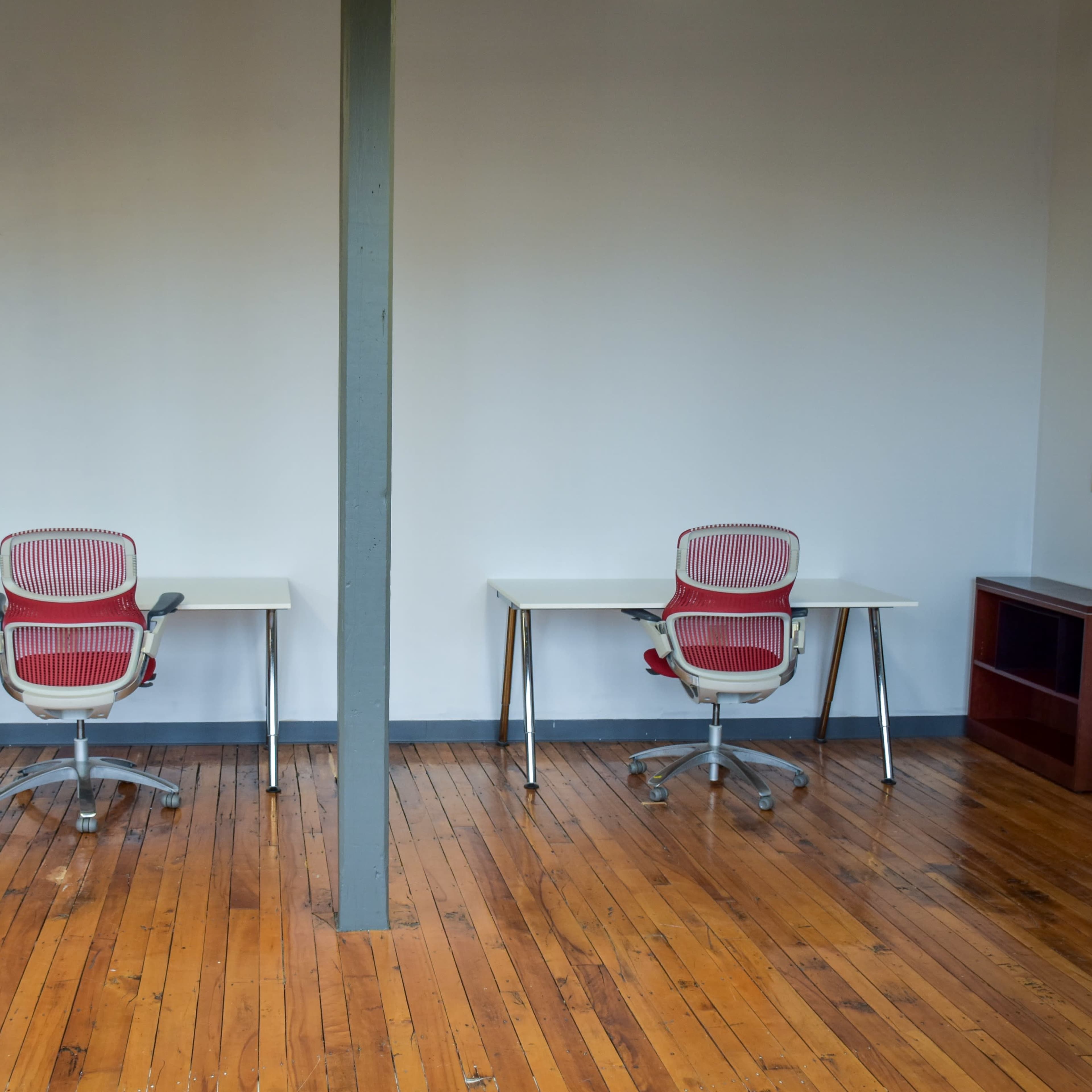 Two empty desks with chairs facing each other in a room with wooden floors and a gray wall.