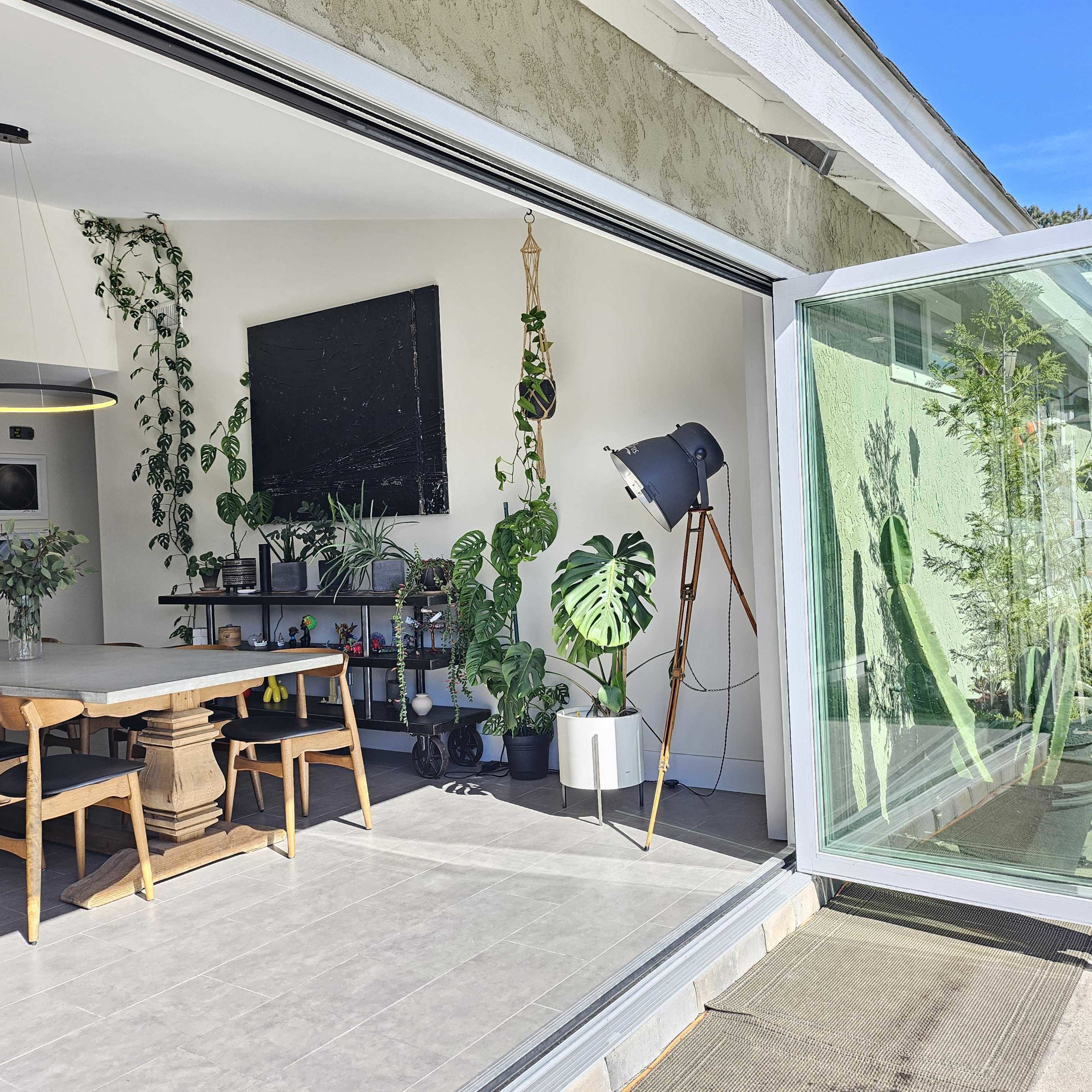 A spacious dining area features a long wooden table surrounded by chairs, large windows open to an outdoor patio, and several indoor plants.