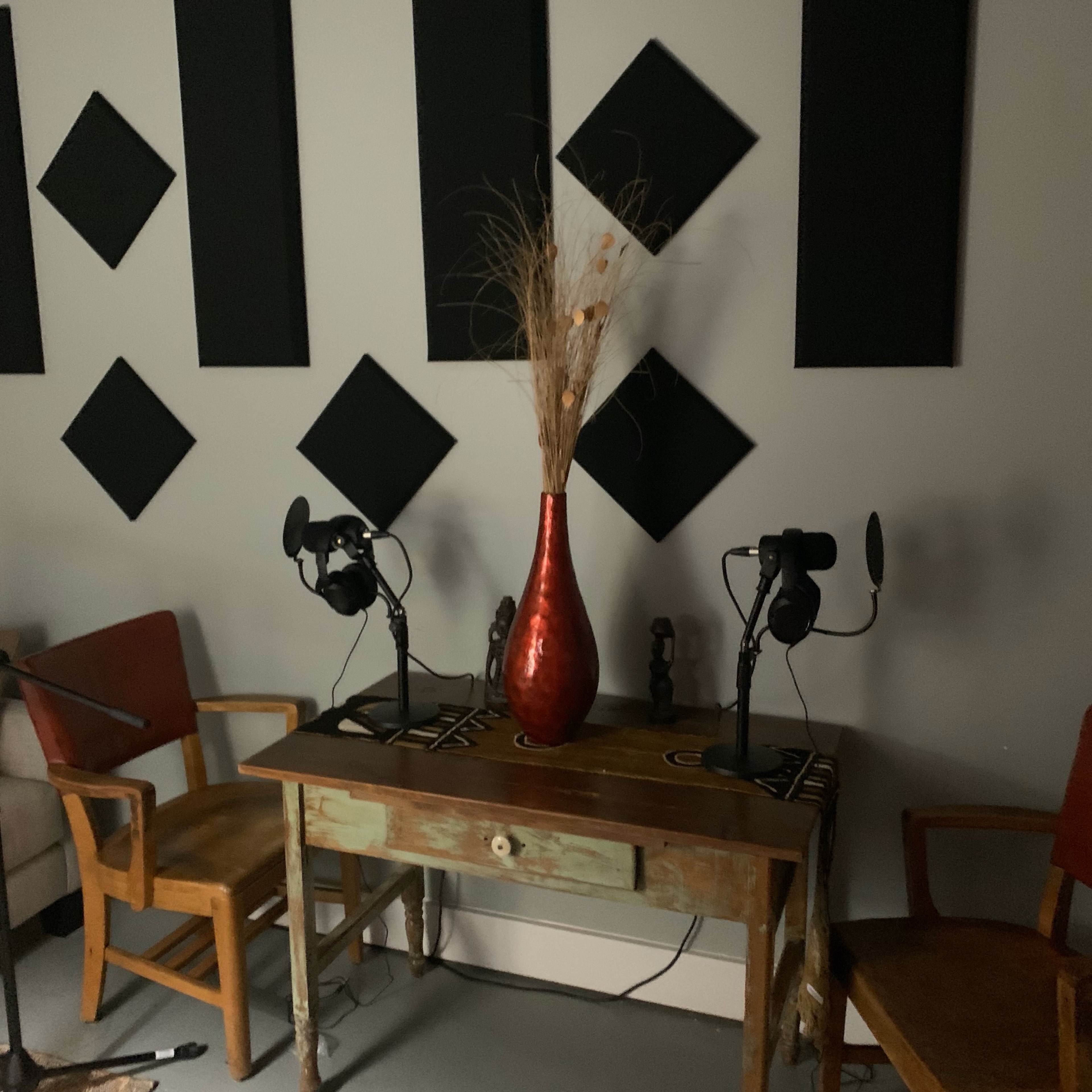 A vintage table with a red vase and dried plants is positioned between two chairs, flanked by two microphones, in a room featuring black diamond-shaped wall panels.