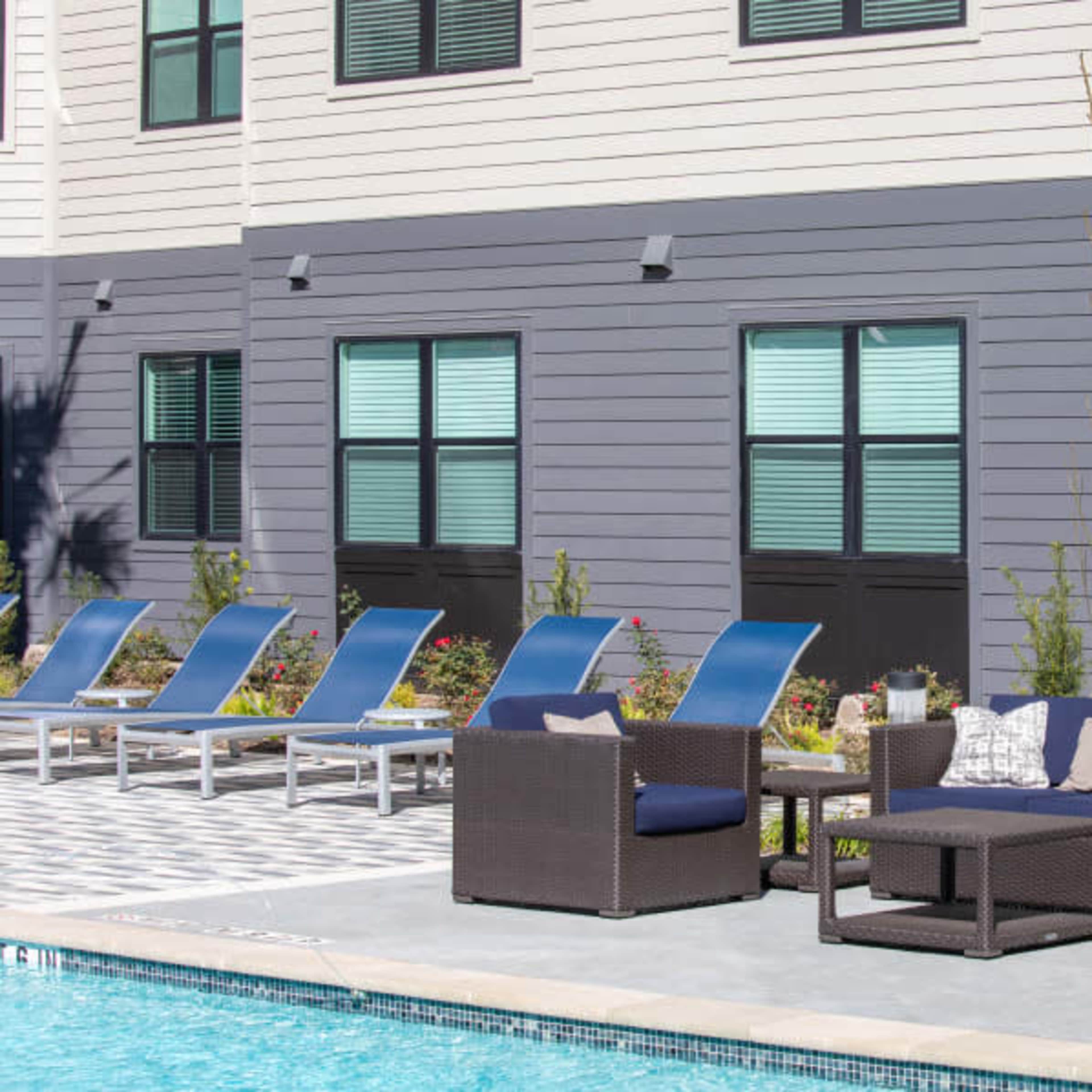 A poolside area features a set of wicker furniture with navy cushions and several lounge chairs arranged along the water's edge.