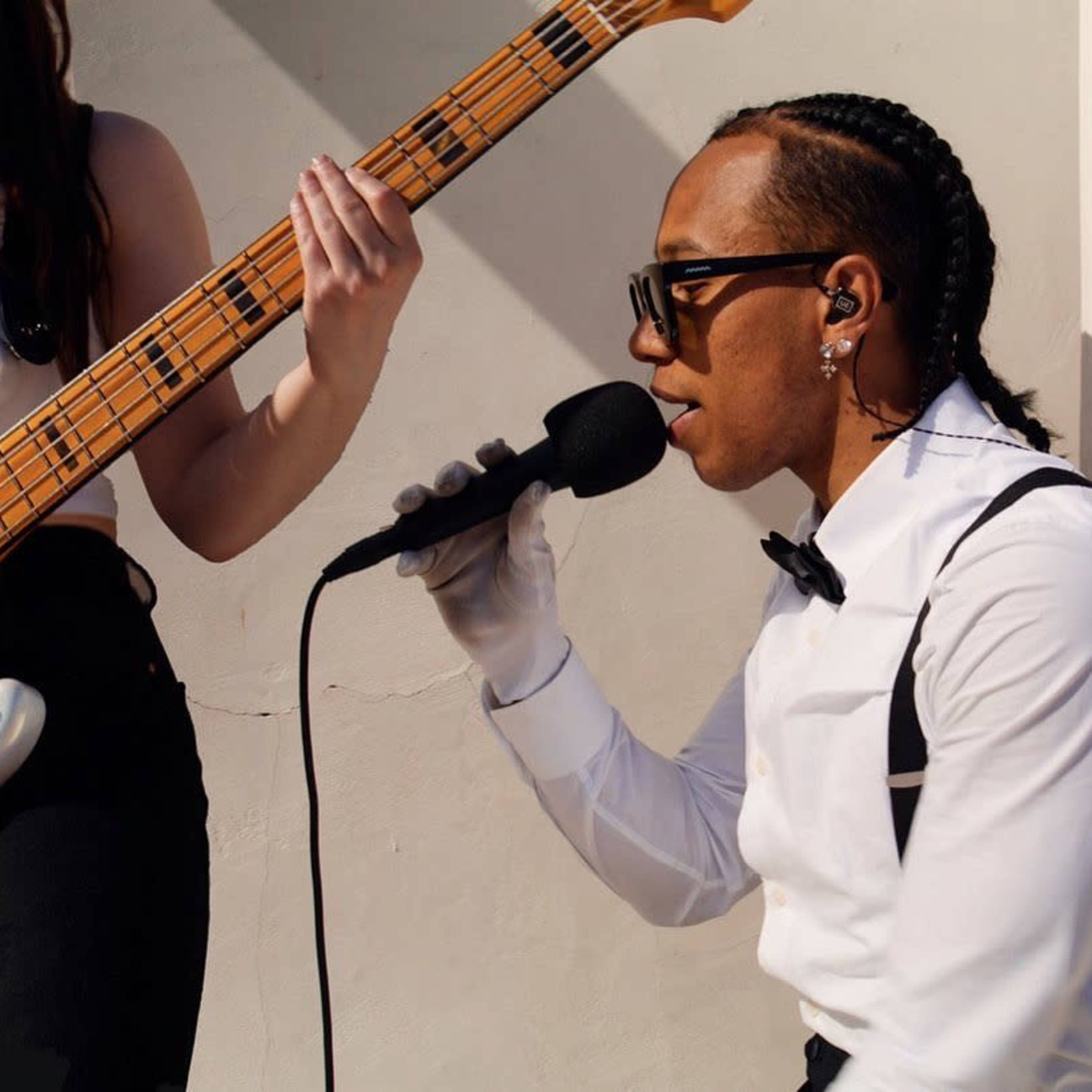 A man in a white shirt and bow tie sings into a microphone while a woman with a bass guitar stands beside him.