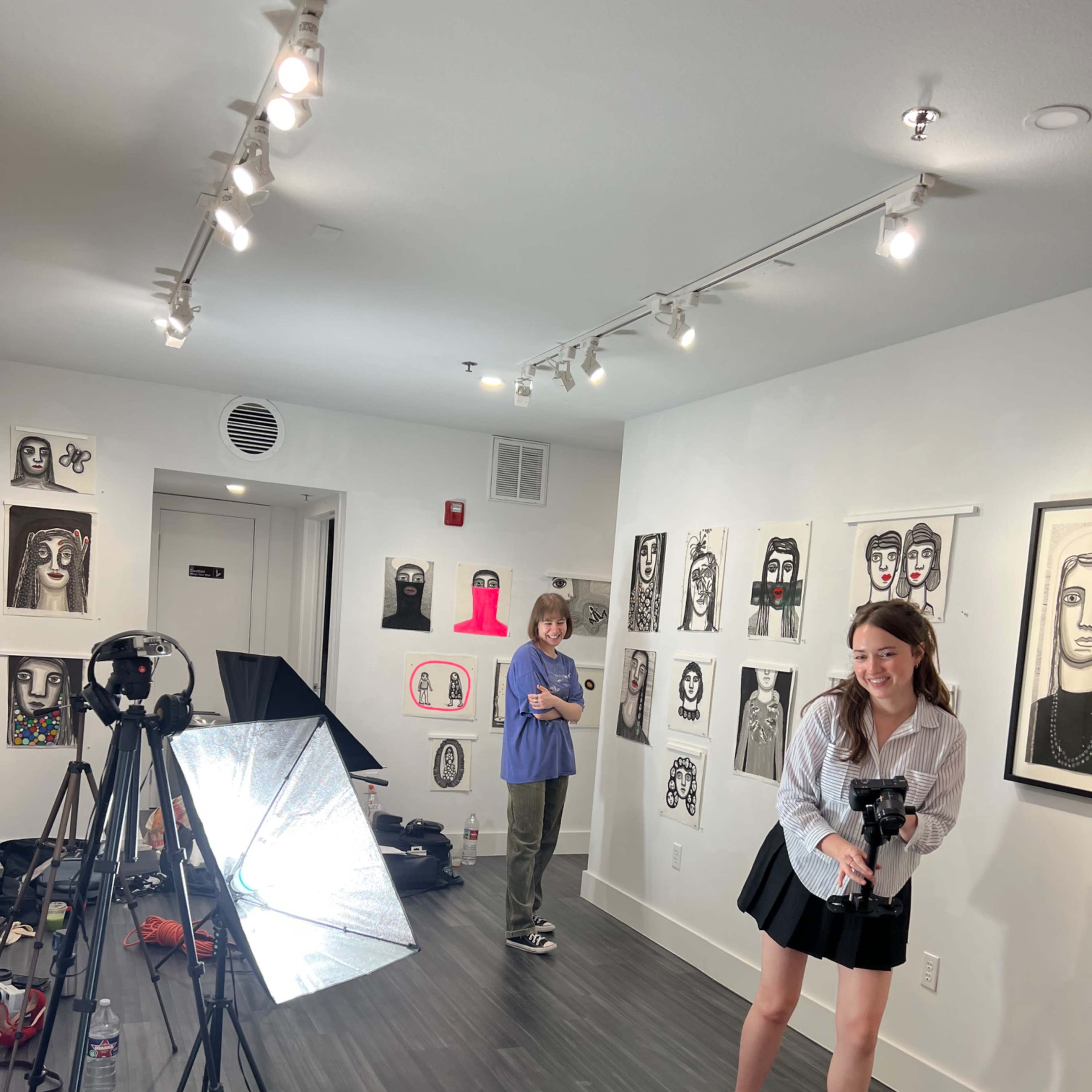 Two people are in an art gallery, with one holding a camera and the other standing near illustrations on the walls while studio lights are set up for a photoshoot.