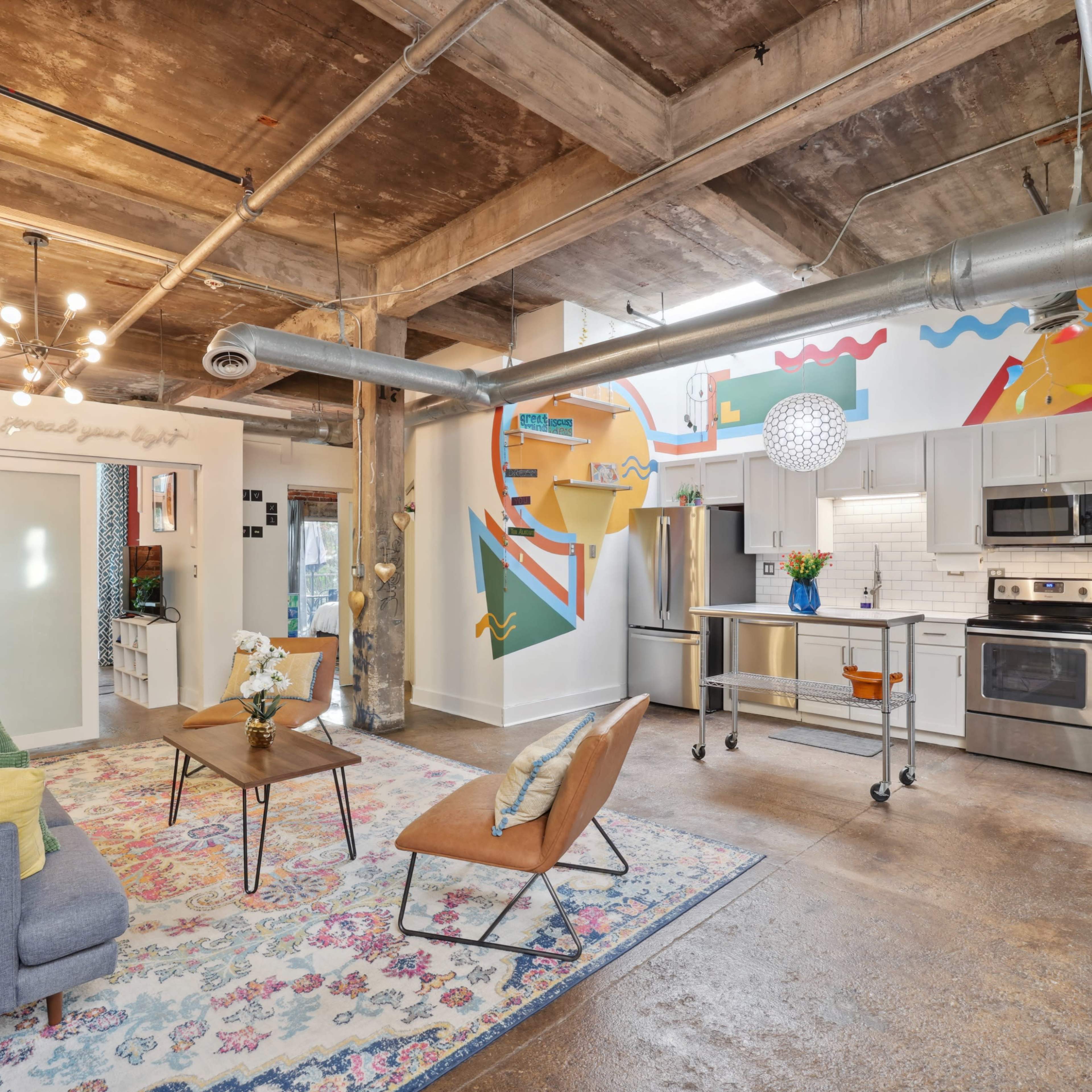 A modern loft-style living space with exposed concrete ceilings, colorful wall murals, and a kitchen area featuring stainless steel appliances.
