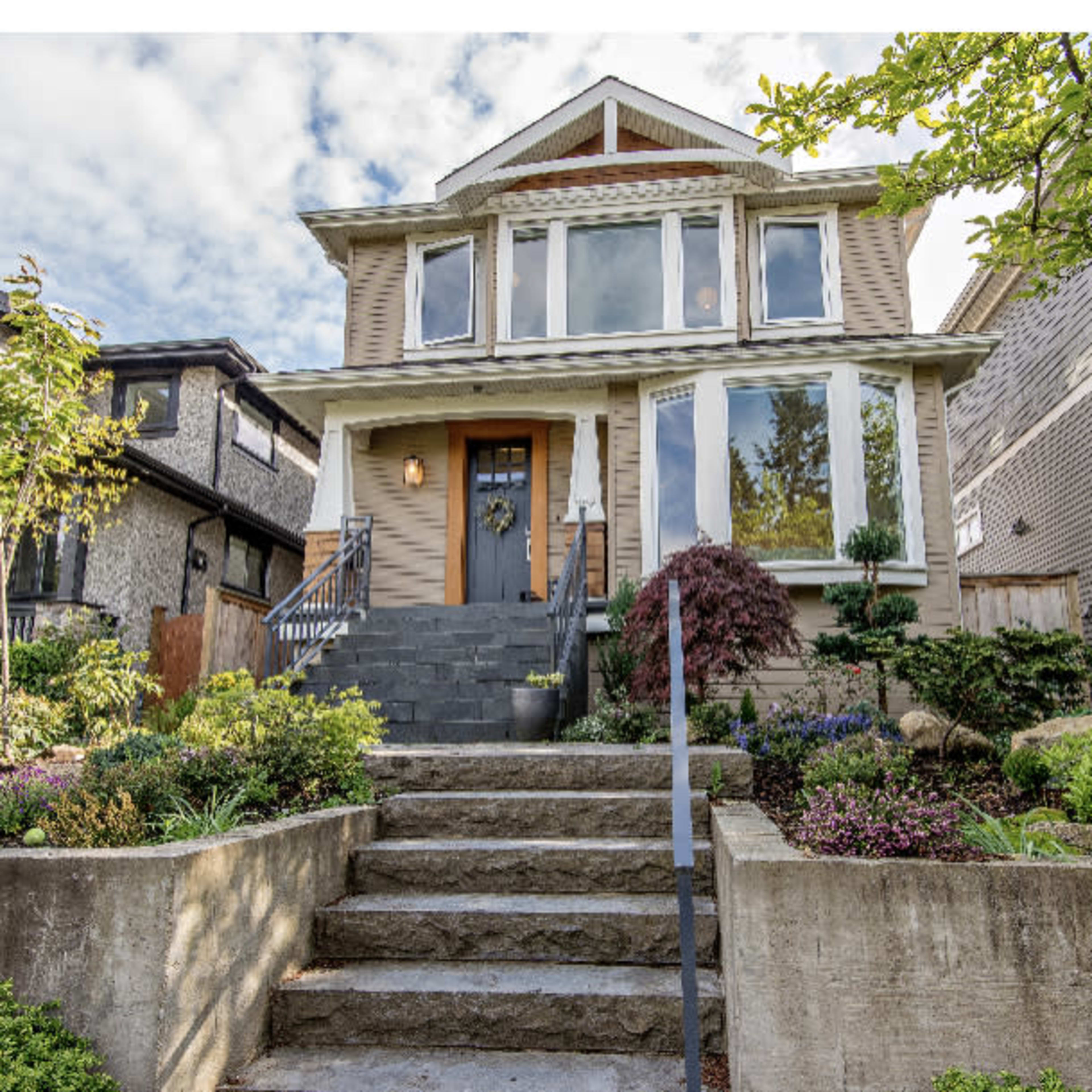 A two-story house features a front porch, landscaped yard with flowers, and stone steps leading up to the entrance.