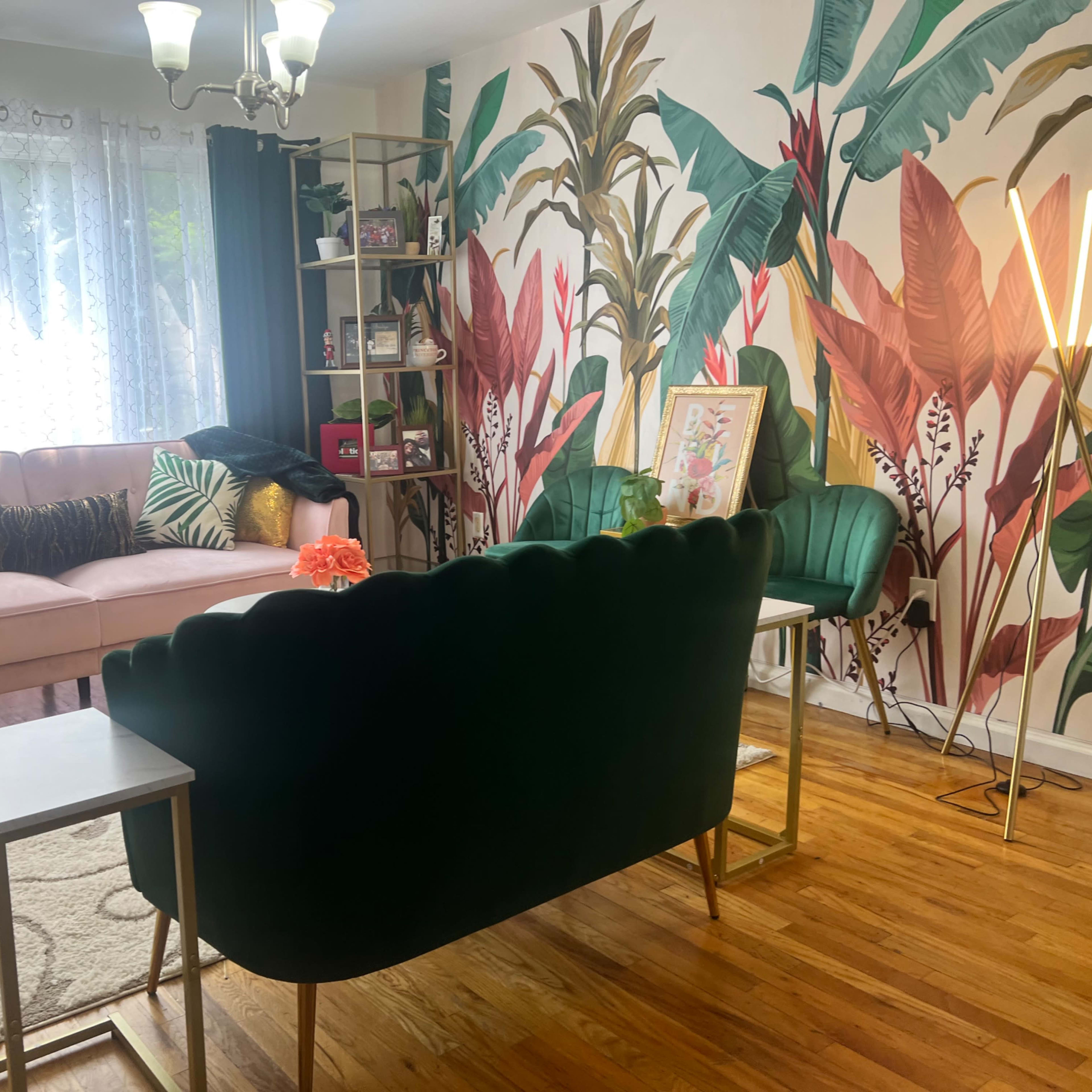 A living room featuring a pink sofa, a green velvet couch, a small white table, and a decorative wall with tropical plants wallpaper.