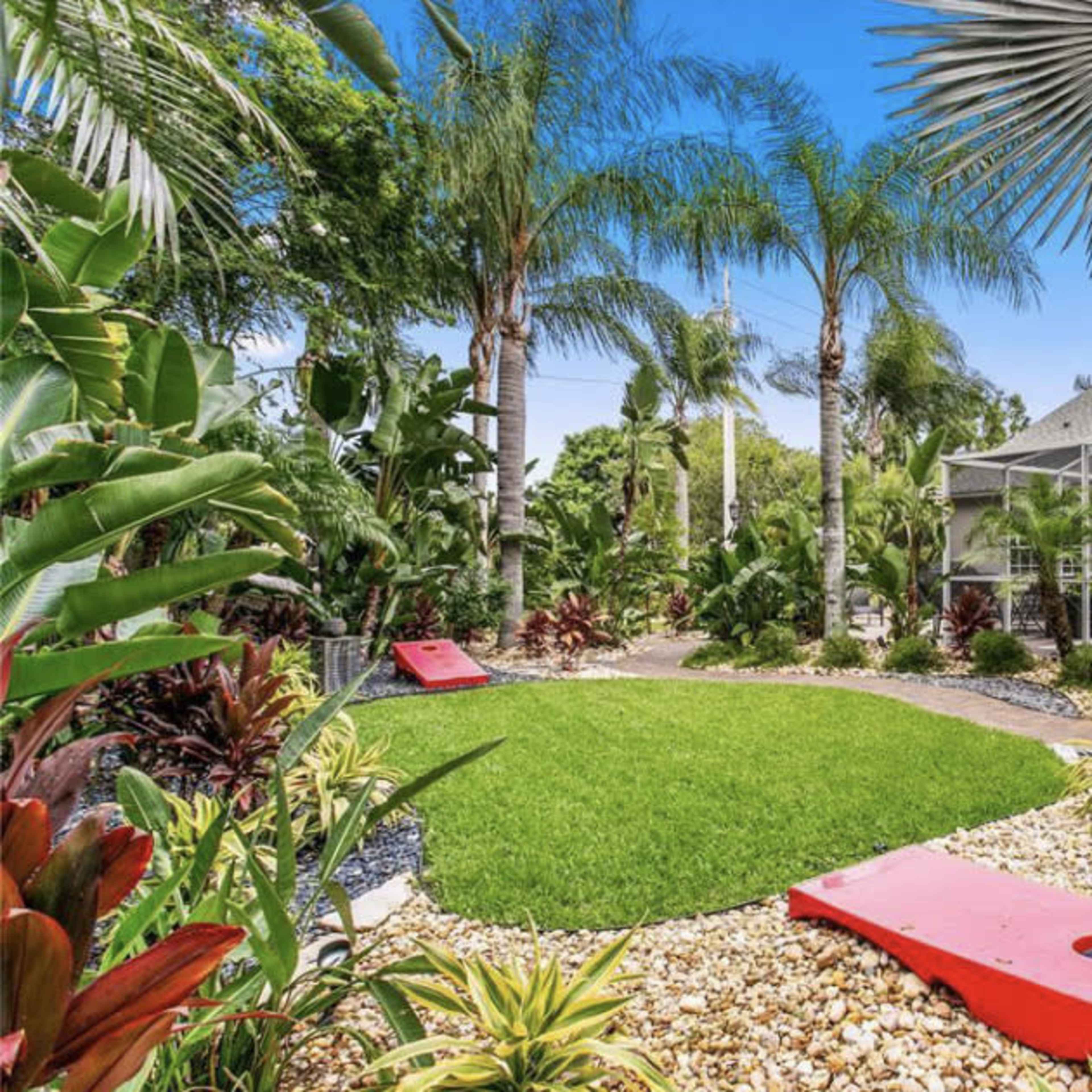 A landscaped backyard with palm trees, colorful plants, a green lawn, and cornhole boards set up on the ground.