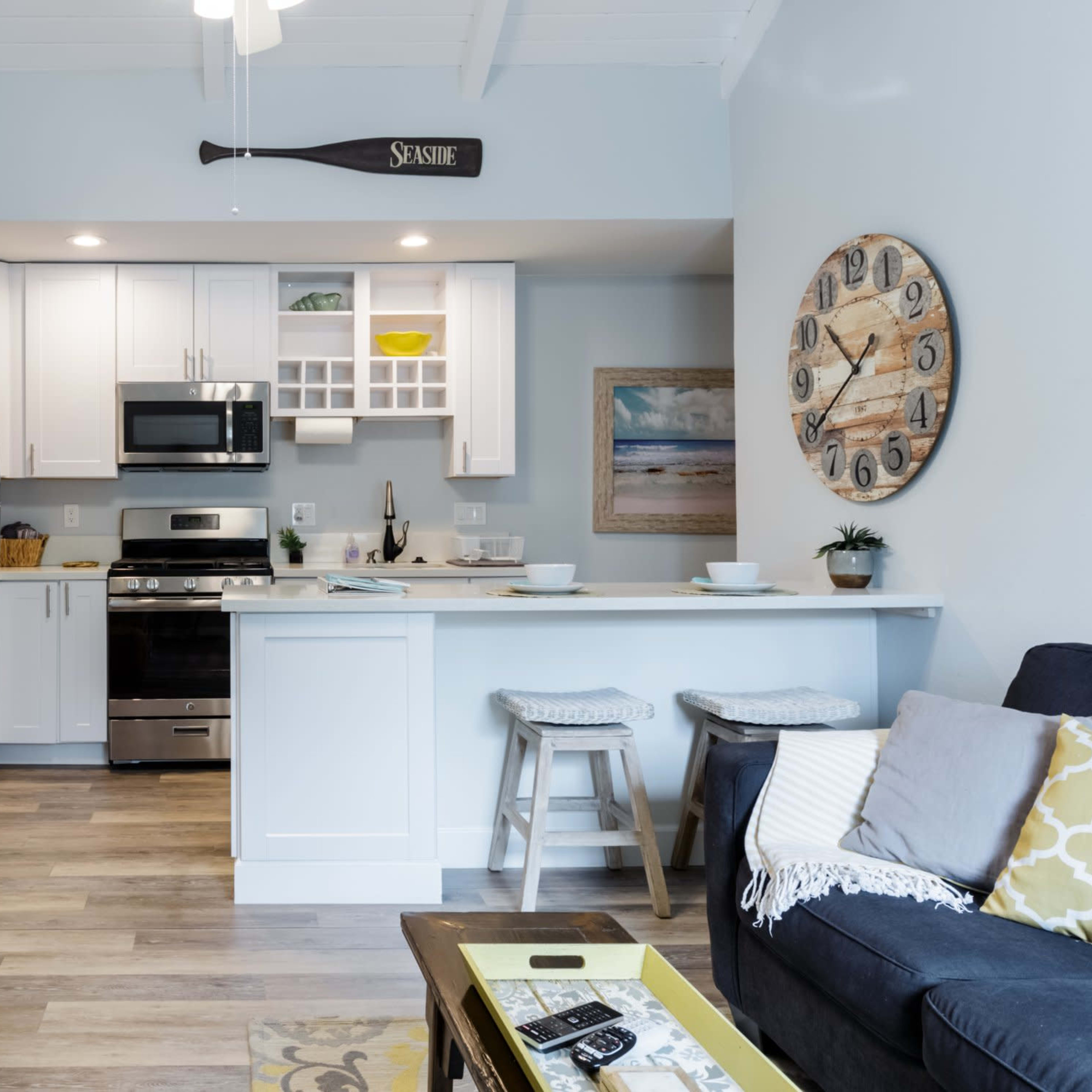 A modern living room and kitchen area with a dark sofa, a wooden coffee table, and white cabinetry, featuring stainless steel appliances and a decorative wall clock.