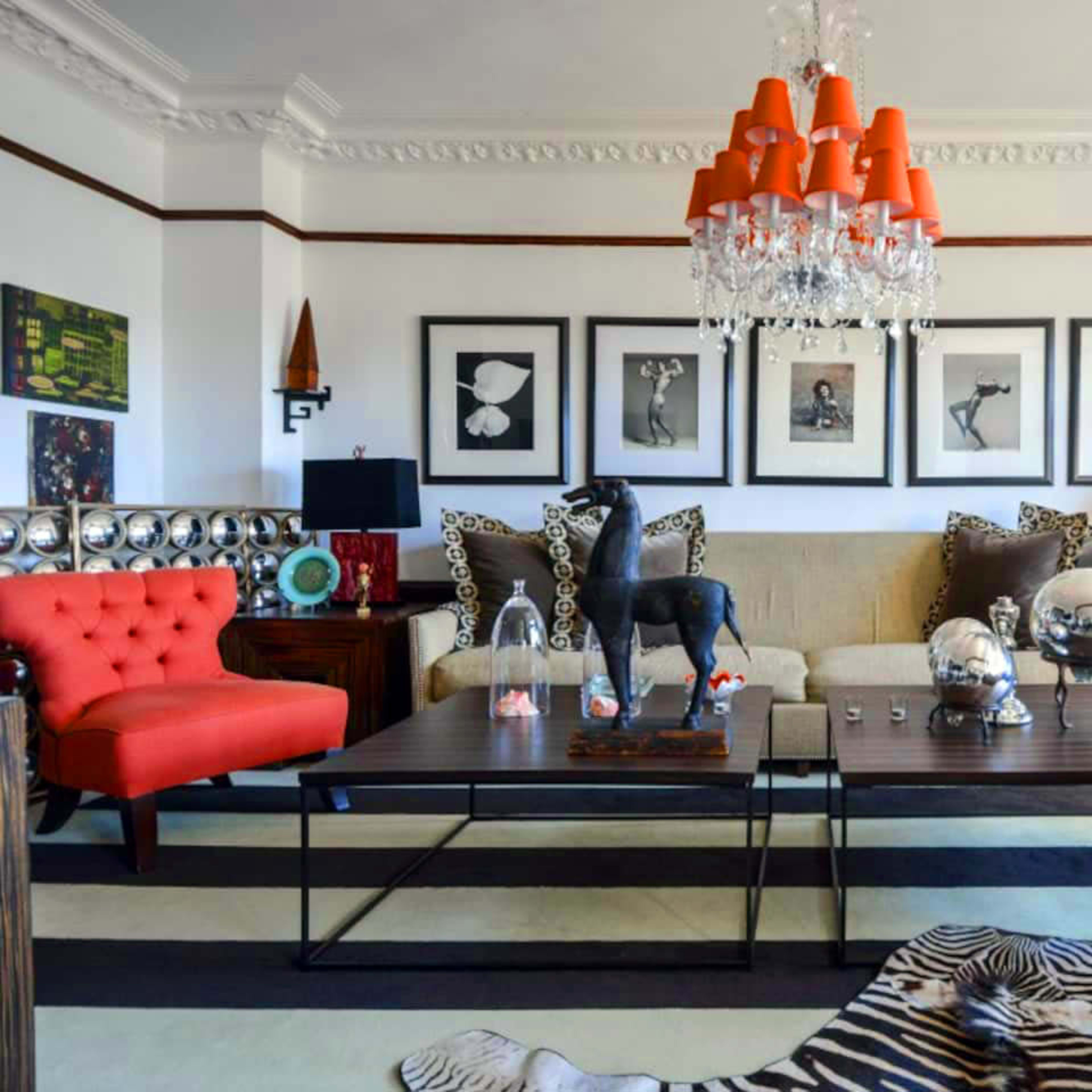 The living room features a mix of bold furniture, including a red tufted sofa and a large coffee table, with artwork and decorative items on display against a backdrop of white walls and patterned rugs.
