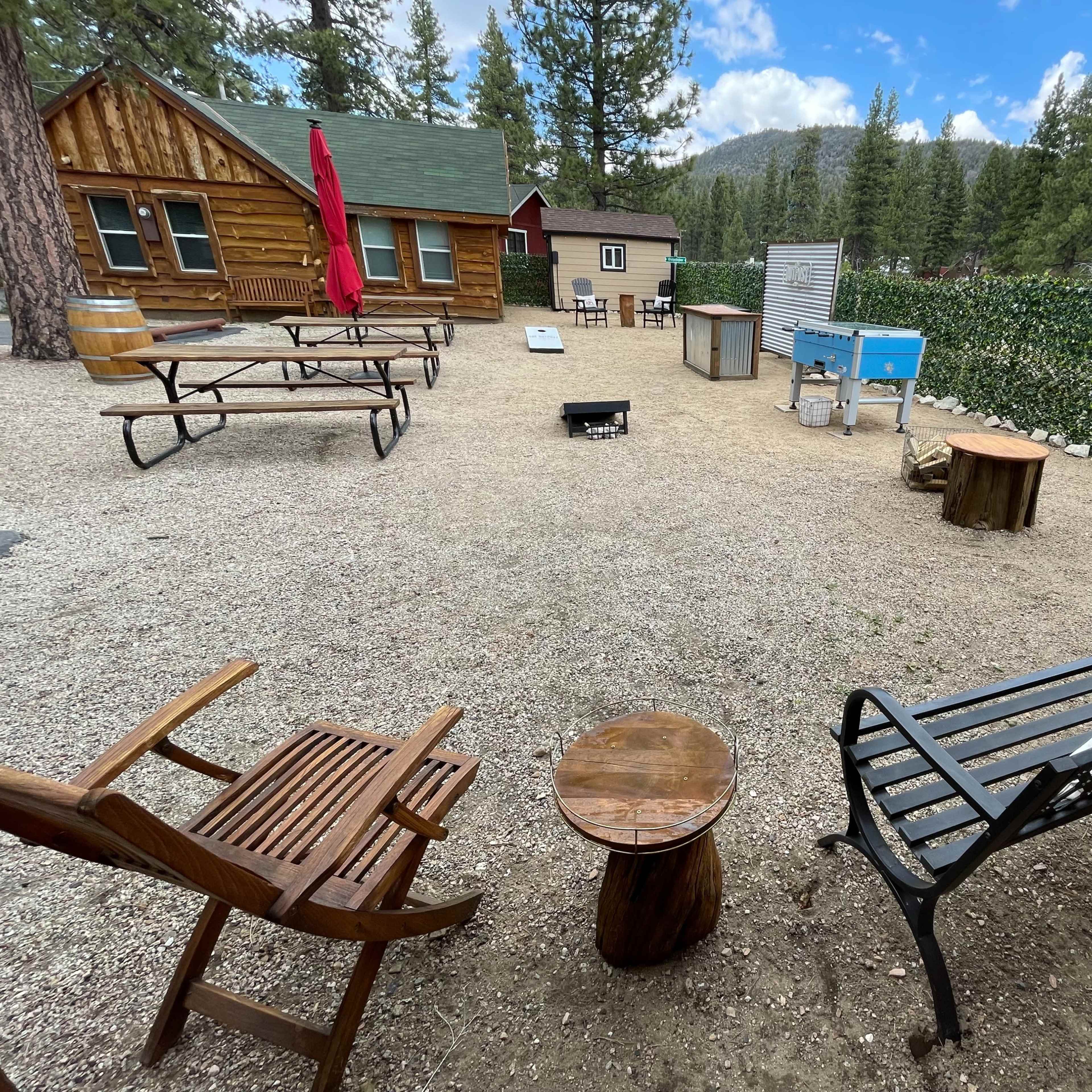 The scene shows a gravel area with wooden seating, picnic tables, and a blue cooler, surrounded by trees and cabins.