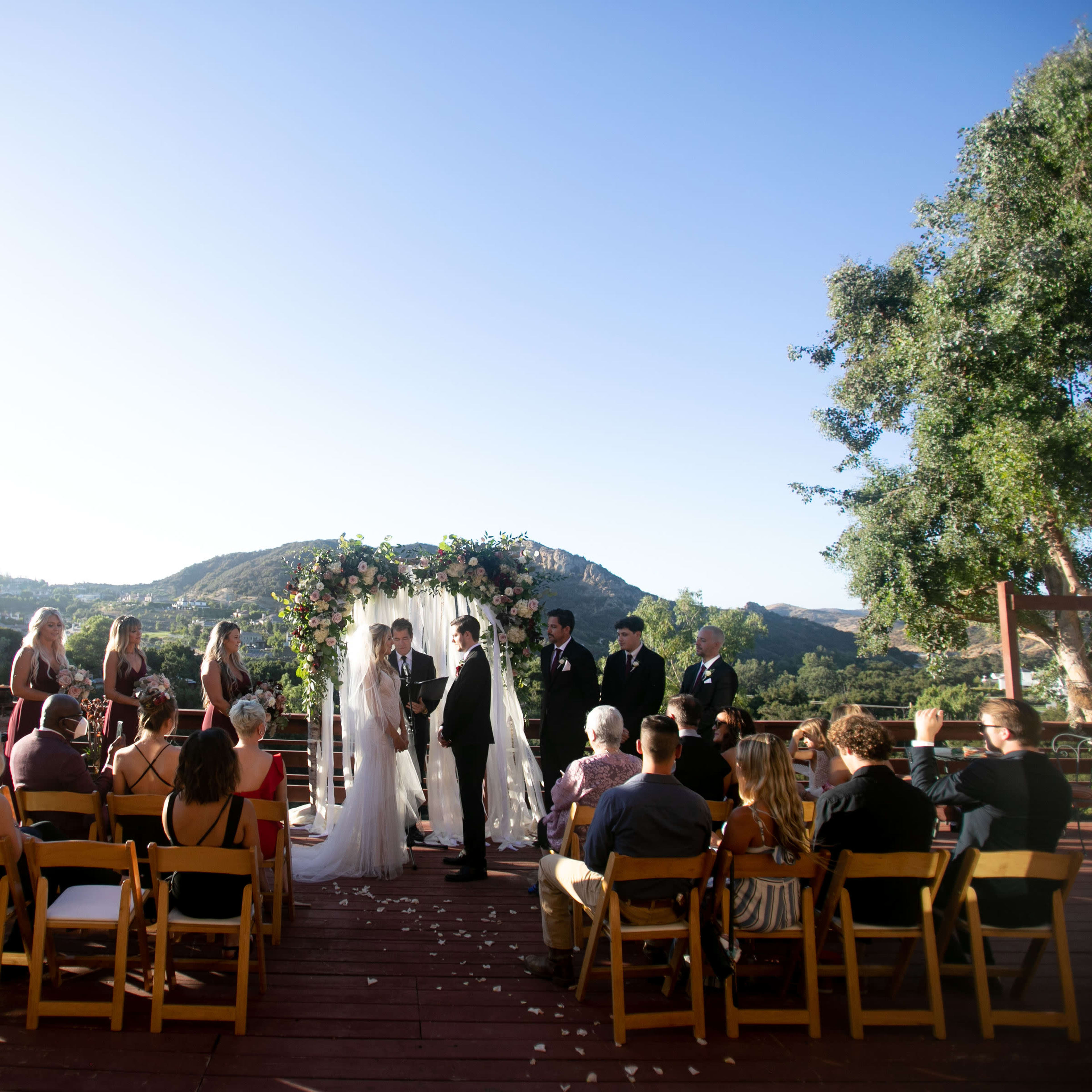 A couple stands at an altar surrounded by guests seated on wooden chairs, with a backdrop of hills and clear blue sky during a wedding ceremony.