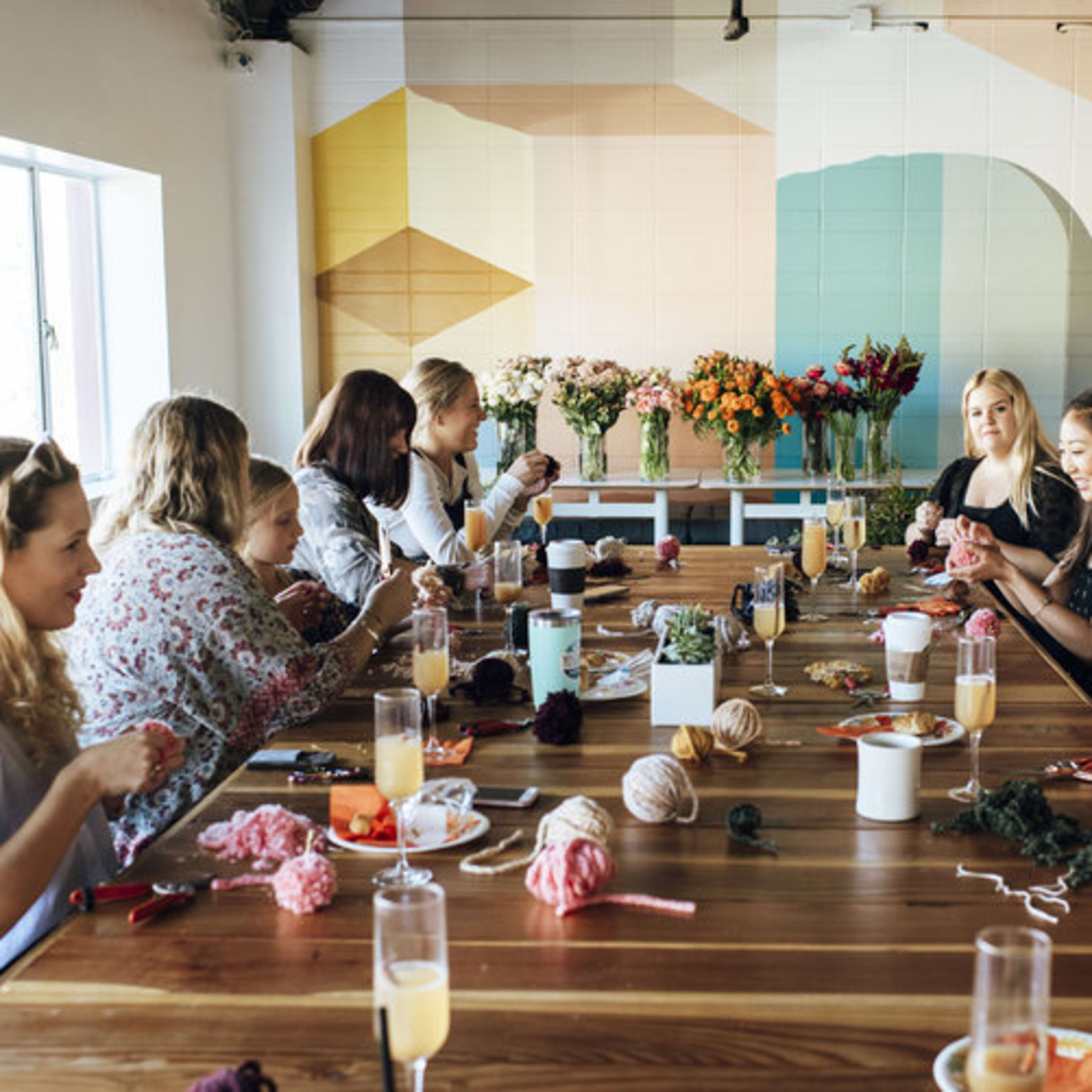 A group of people are seated around a long wooden table, engaged in a crafting activity with yarn and various materials, while colorful flowers are displayed in vases nearby.