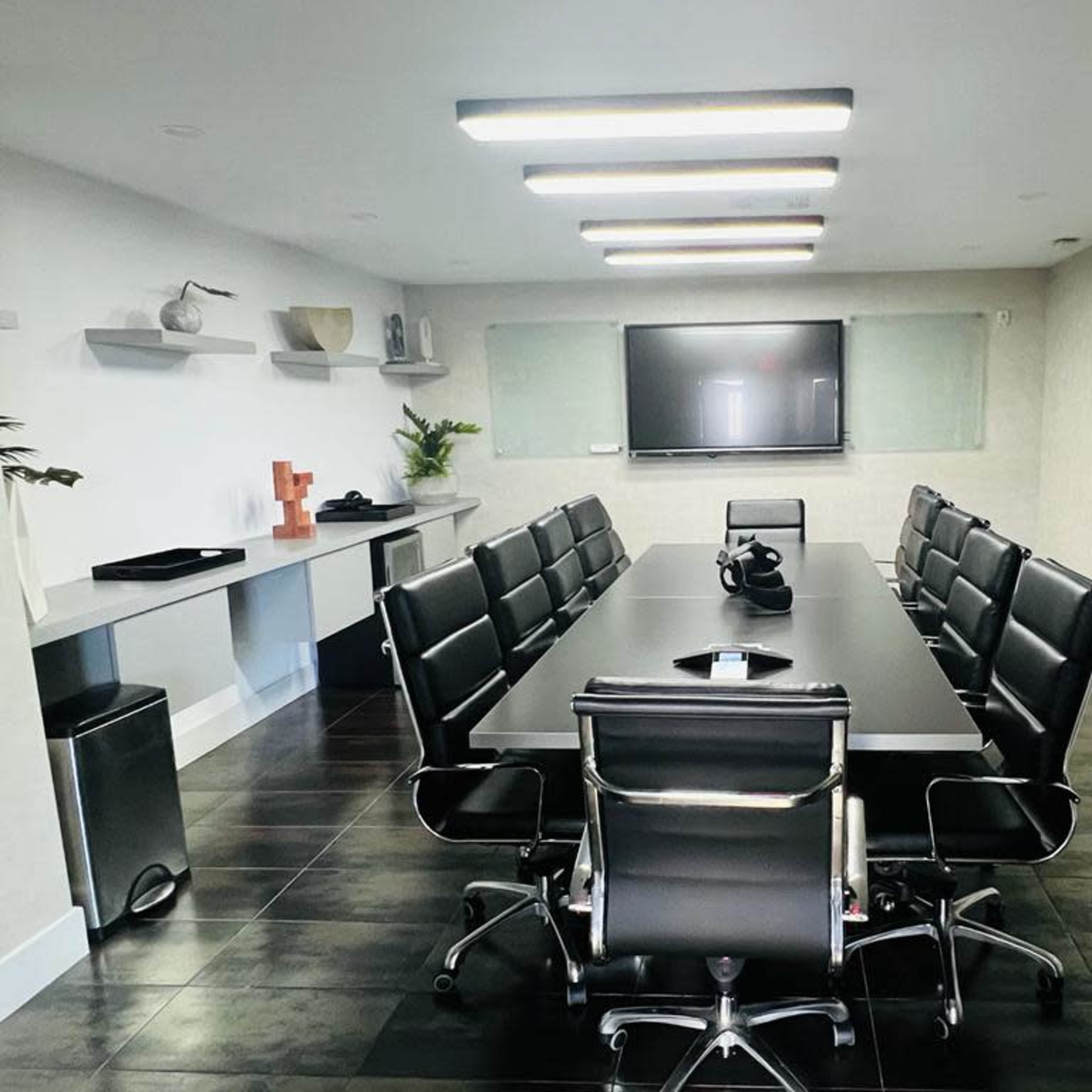 A conference room features a large rectangular table surrounded by black leather chairs, with a television mounted on the wall and a painting beside it.