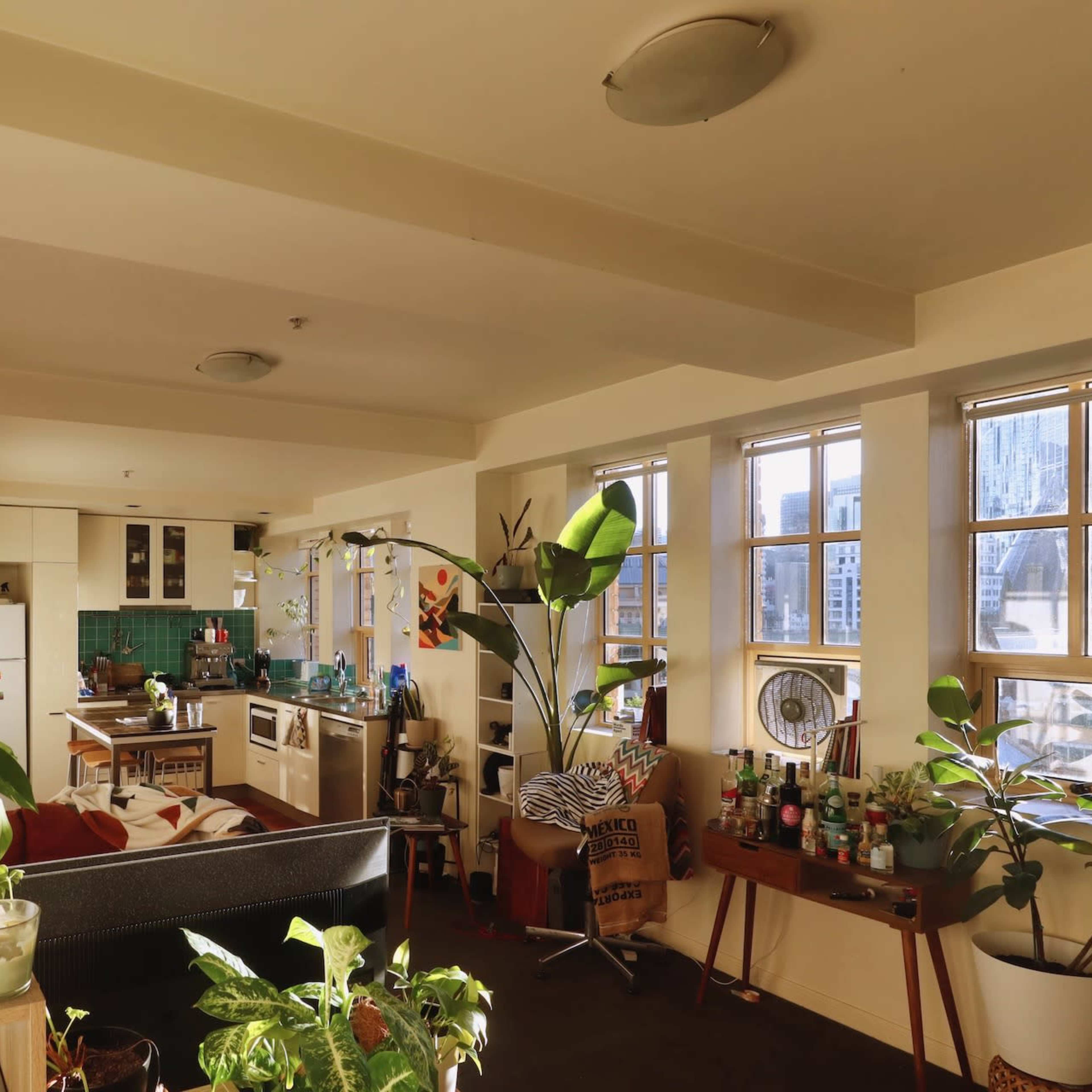 A bright, open-plan living space features a kitchen, a seating area with plants, and large windows showcasing a city view.