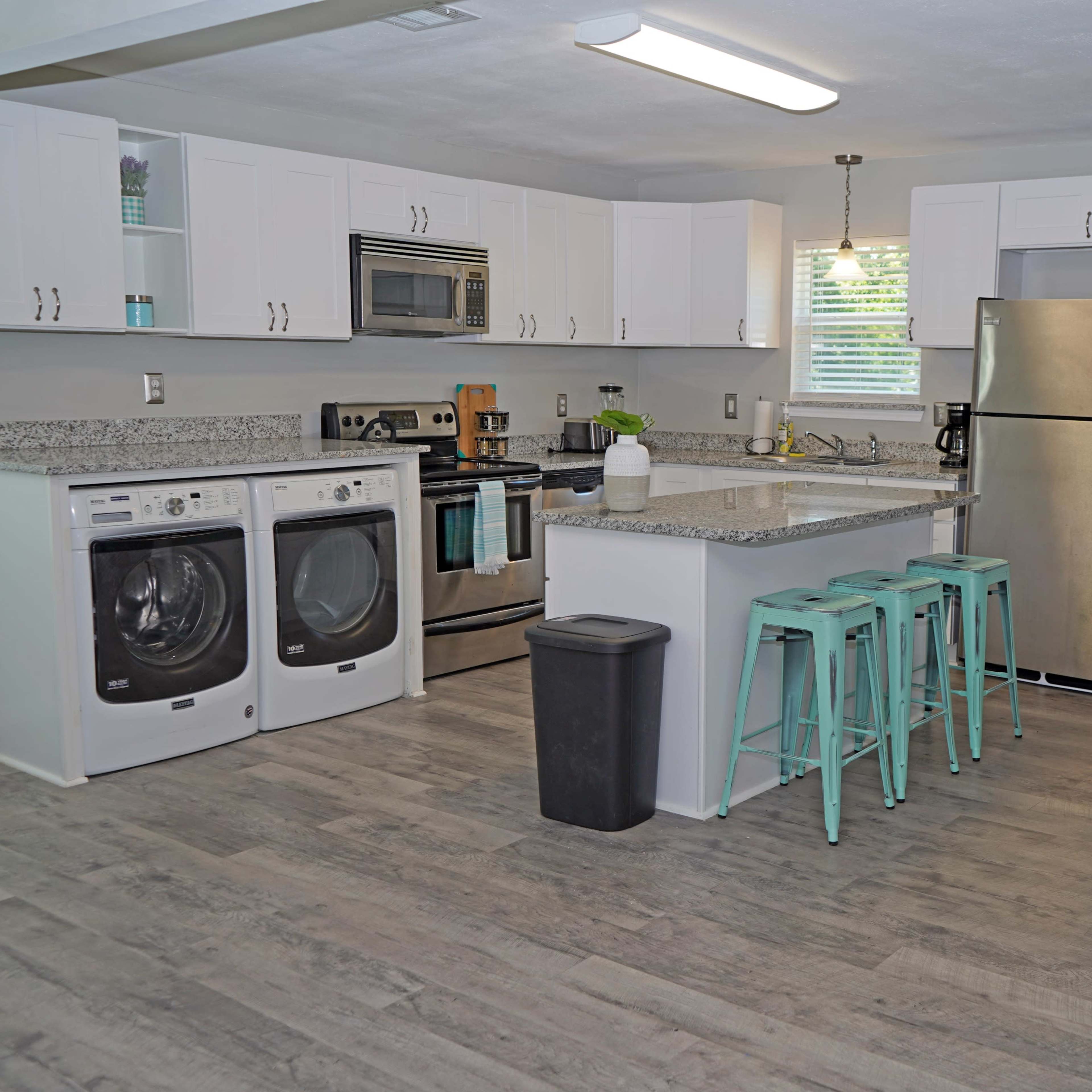 A modern kitchen features a washer and dryer, stainless steel appliances, a centrally located island with turquoise bar stools, and light-colored cabinetry.