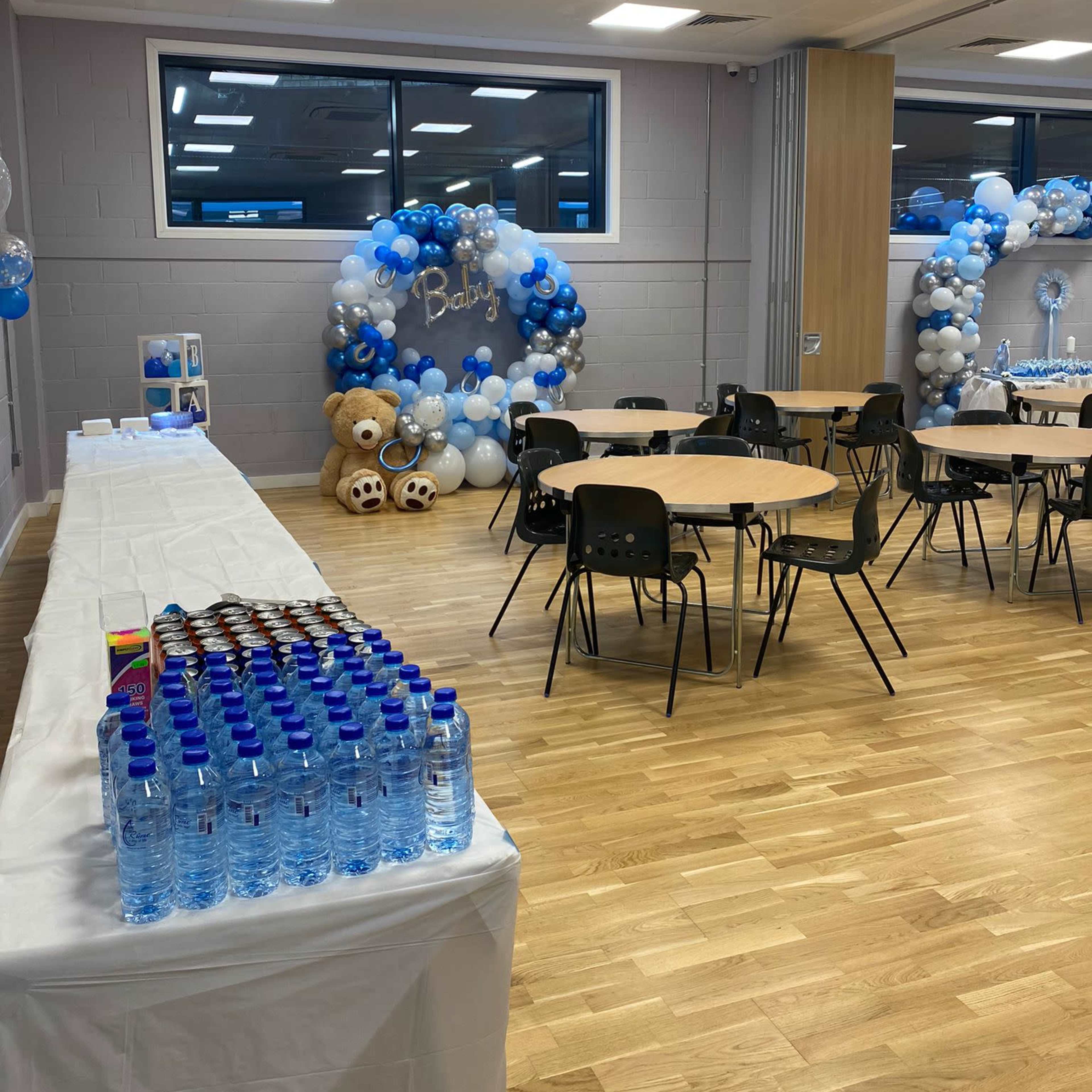 The room is set up for a baby shower, featuring tables with chairs, decorative balloons, bears, and a long table with bottled water.