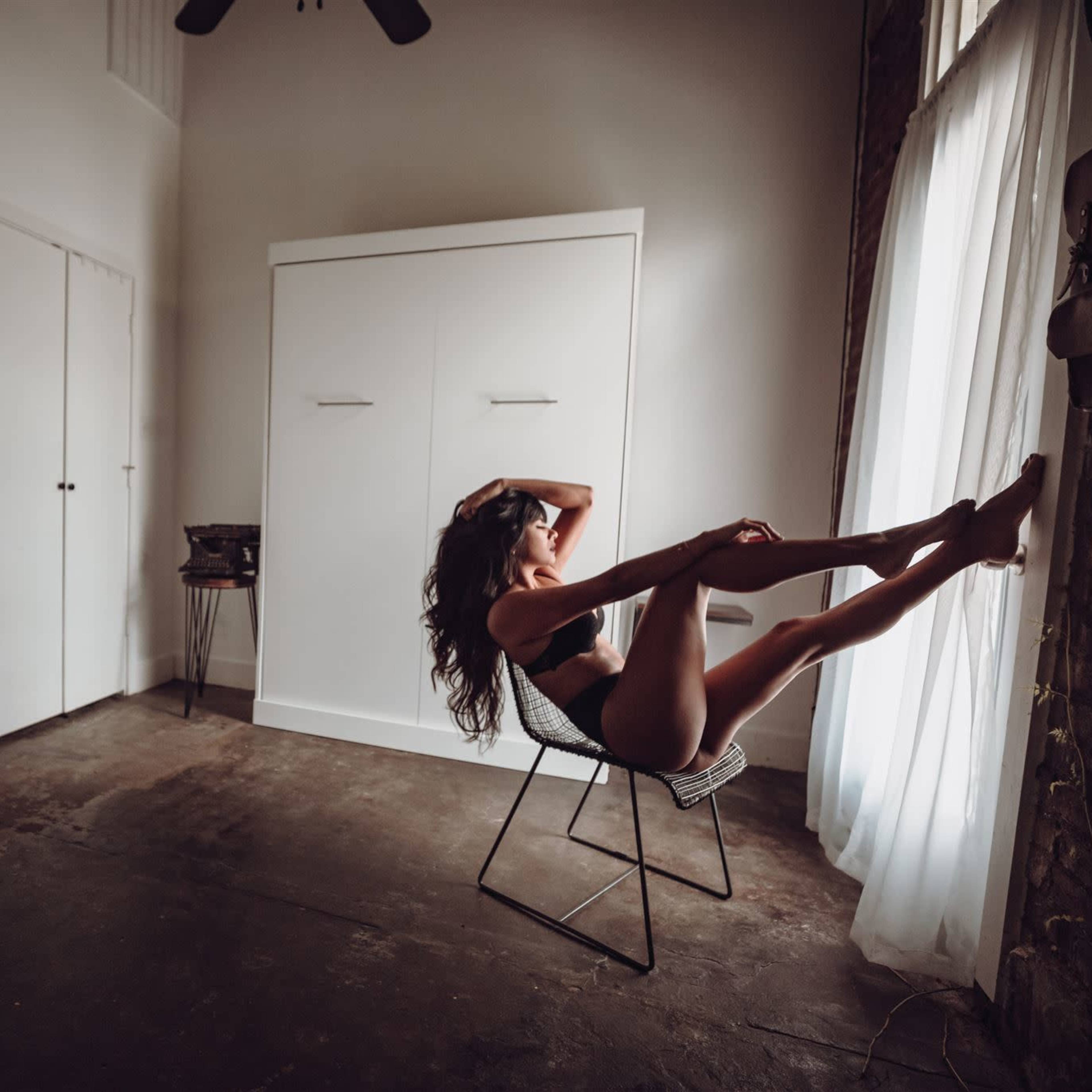 A person sits in a chair by a window in a sparsely furnished room with a concrete floor.