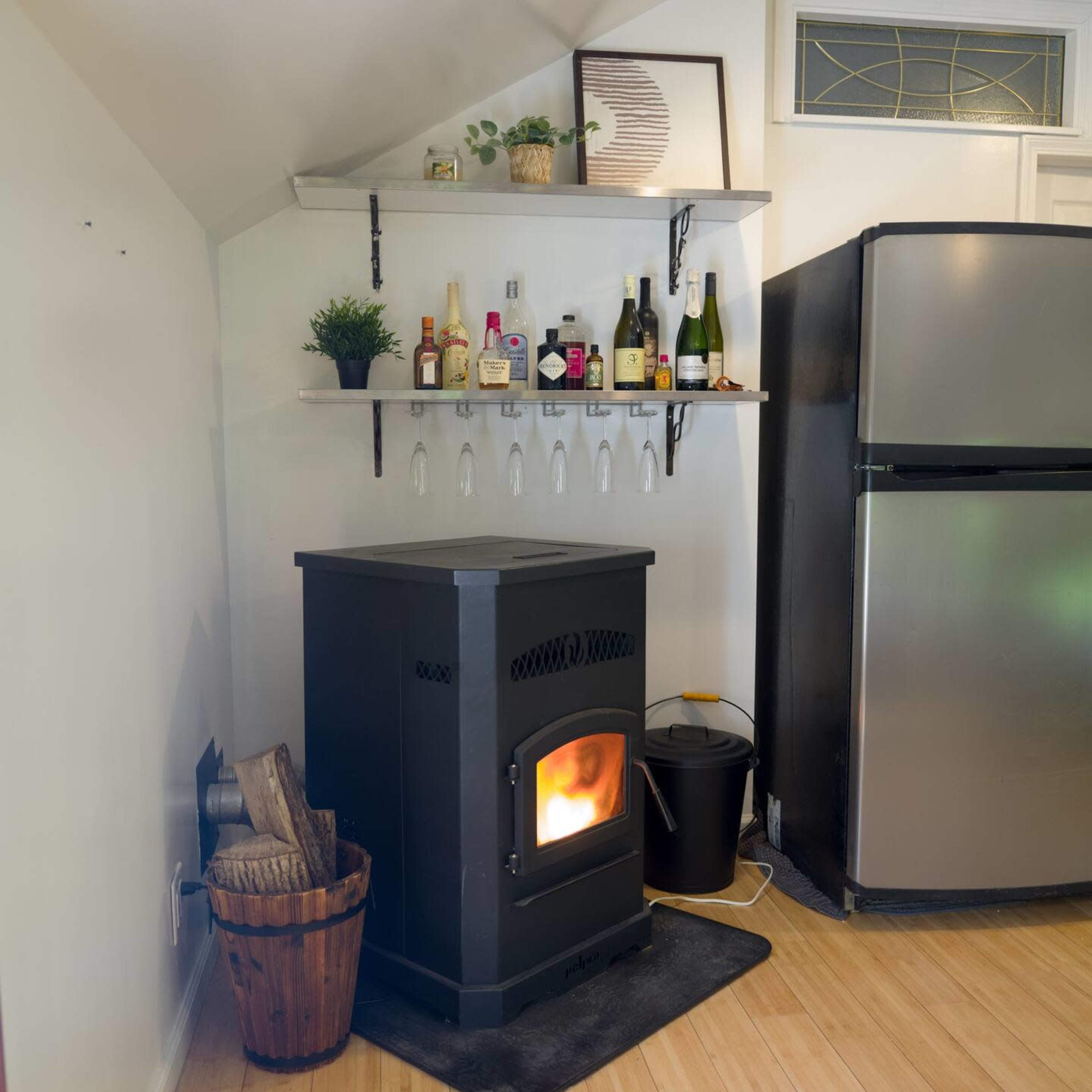 A black stove with an orange flame is positioned near a small shelving unit displaying various bottles, next to a stainless steel refrigerator in a light-colored kitchen.