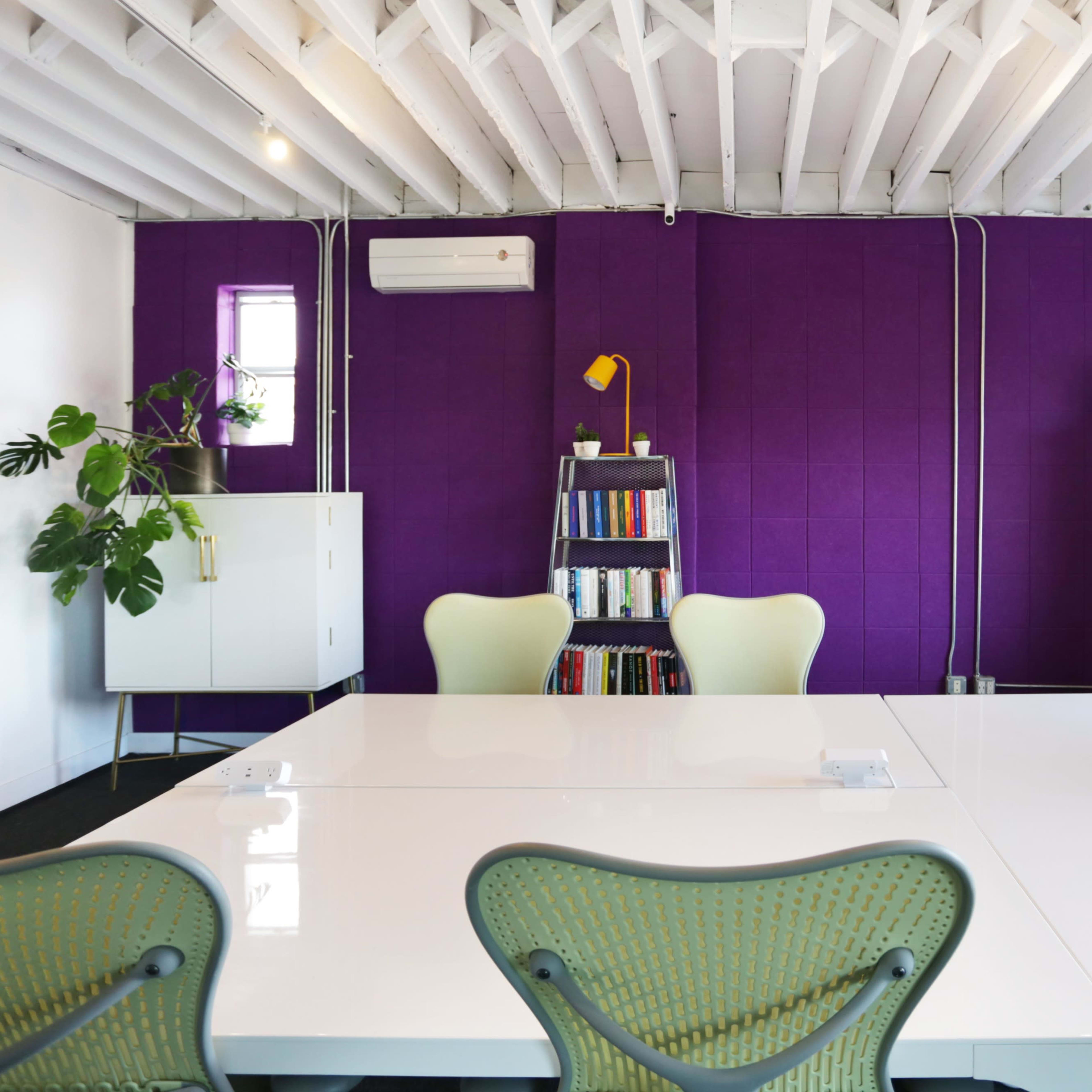 A bright meeting room features a white table, green chairs, a purple accent wall, and shelves with books and plants.