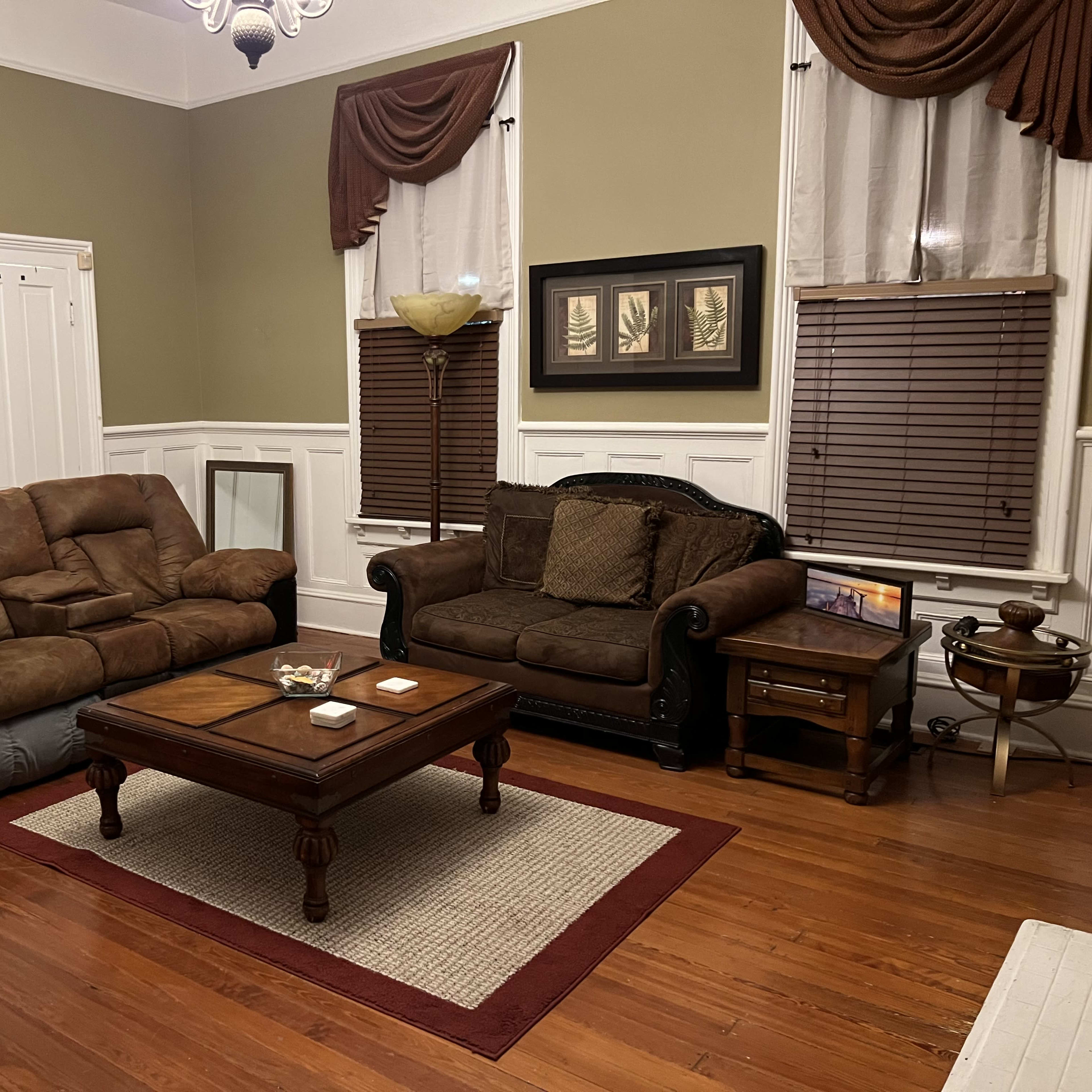 A living room with two sofas, a coffee table, and wooden flooring, framed by brown window treatments and light-colored walls.
