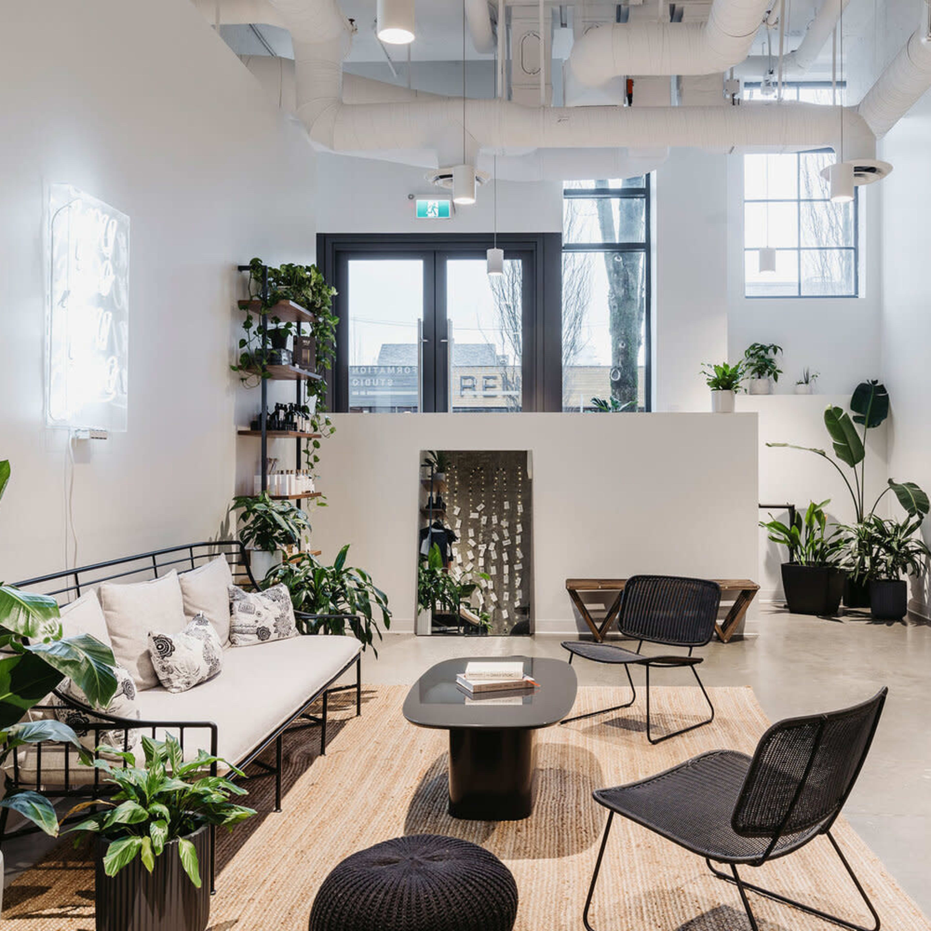 A modern lounge area featuring a sofa, black chairs, a coffee table, and numerous plants, with large windows allowing natural light to enter.