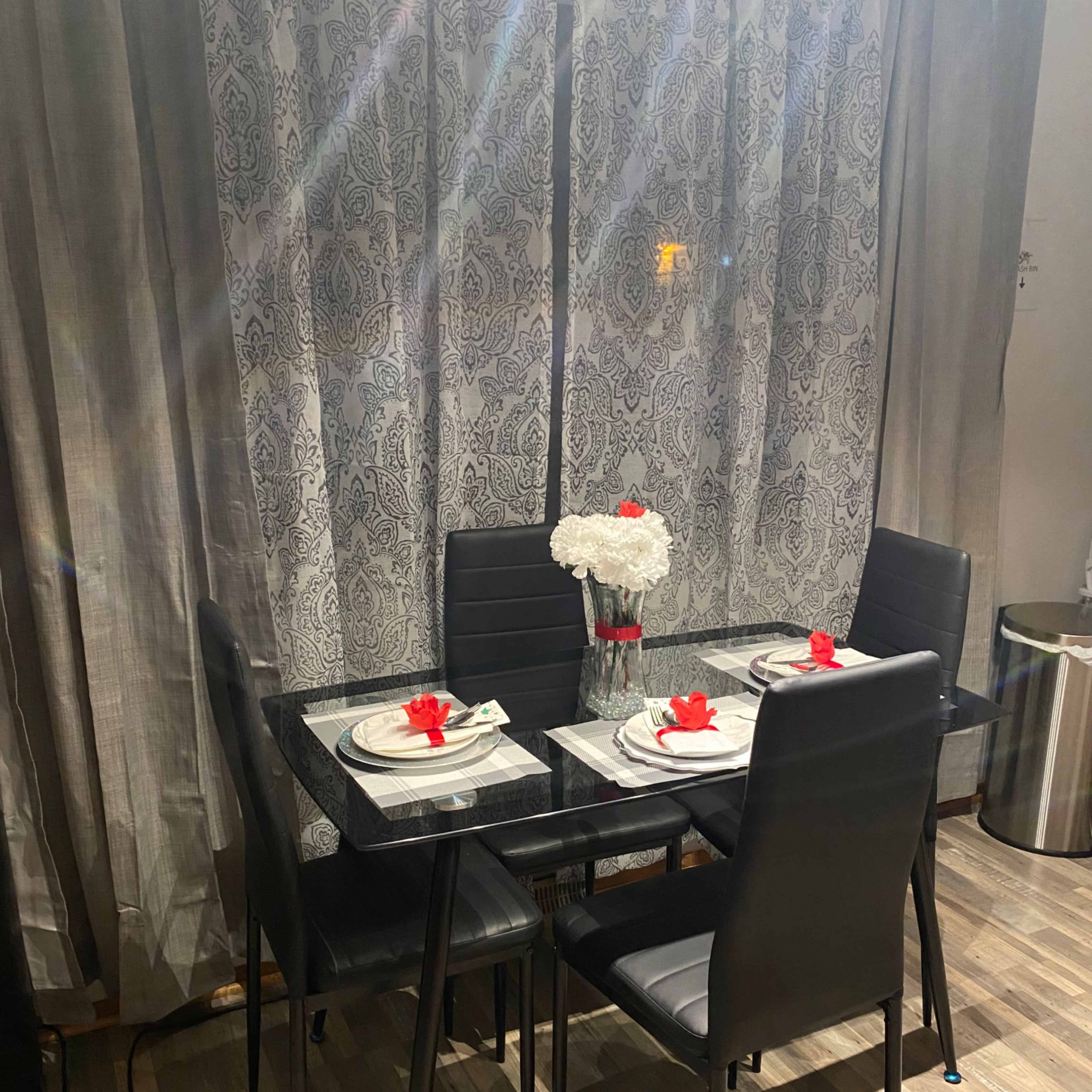 A glass-topped dining table is set with plates and red flowers in a vase, surrounded by four black chairs in front of patterned curtains.