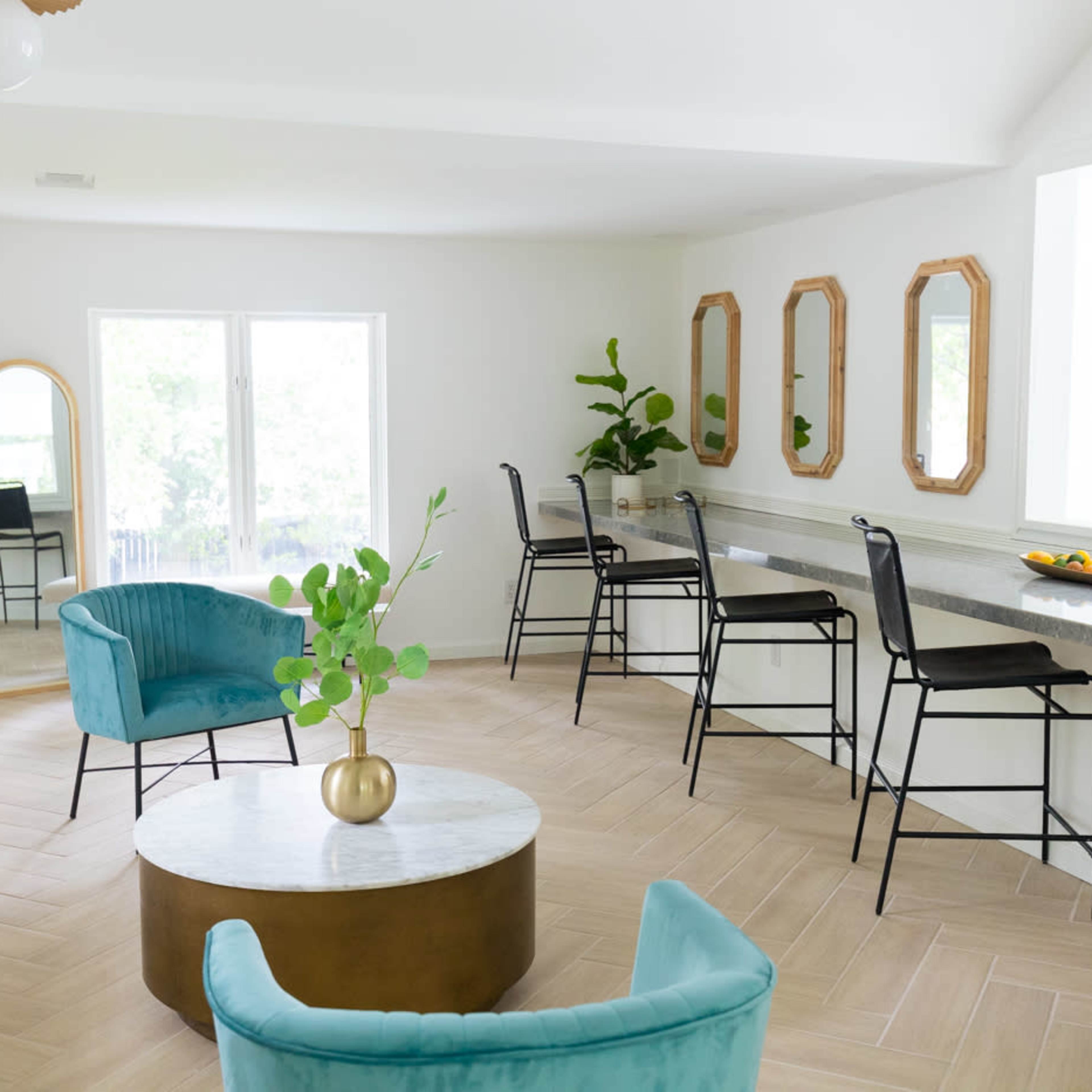 A bright, modern interior features a seating area with two blue chairs, a round coffee table, and a long counter with black chairs and mirrors on the walls.