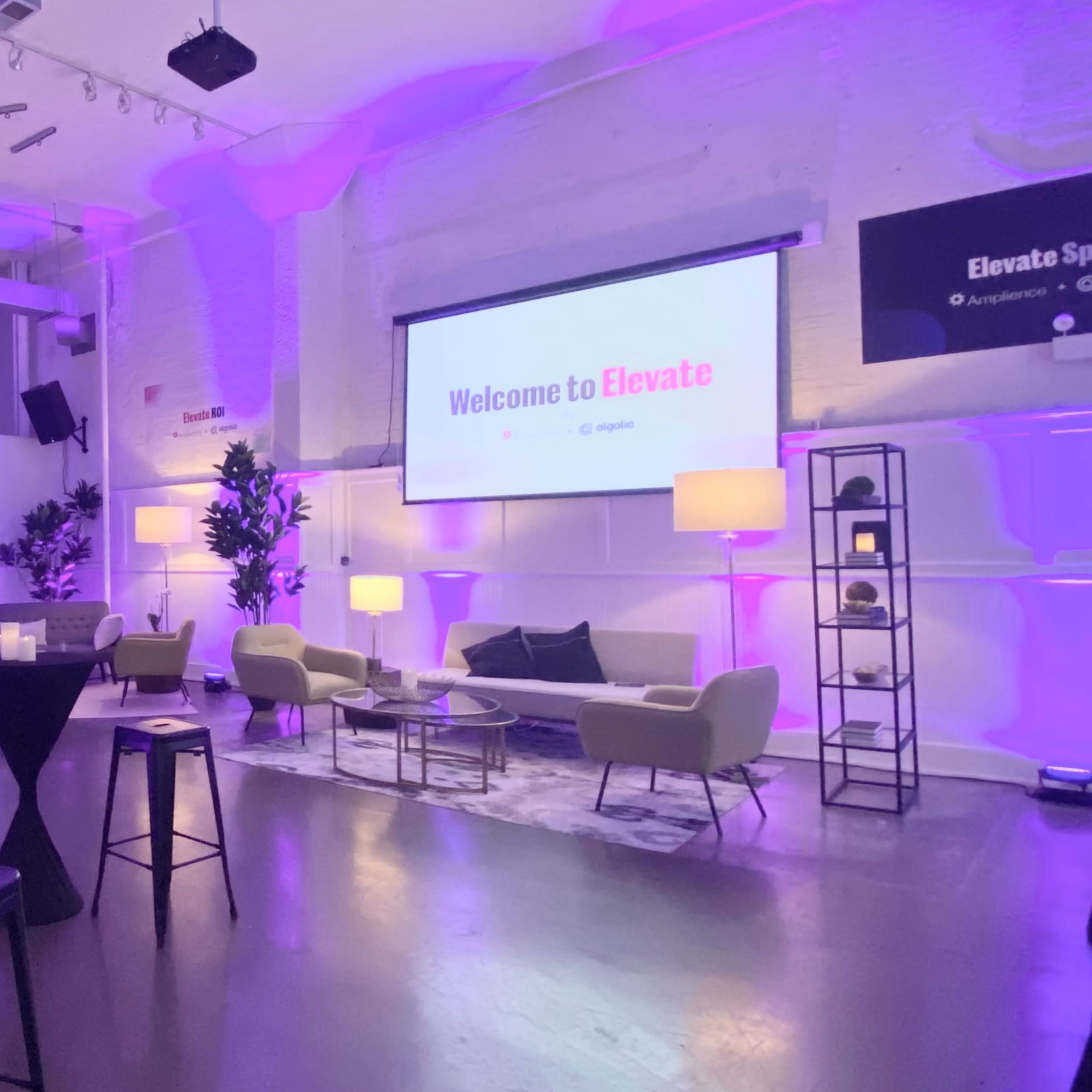 A well-lit event space features seating arrangements, decorative plants, and a large screen displaying "Welcome to Elevate."