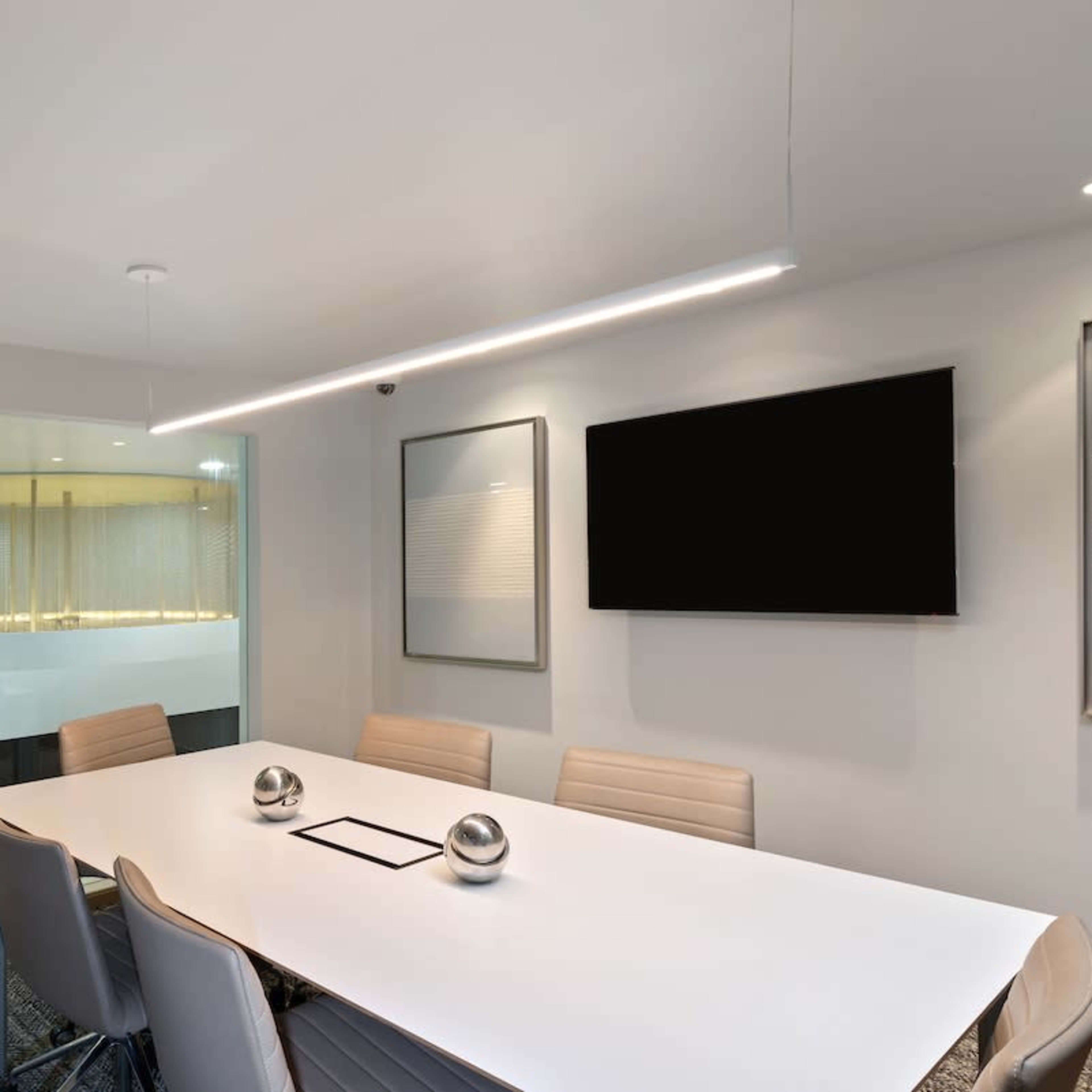 A modern conference room features a large white table surrounded by stylish chairs, with a glass wall leading to an adjacent space and a wall-mounted television.